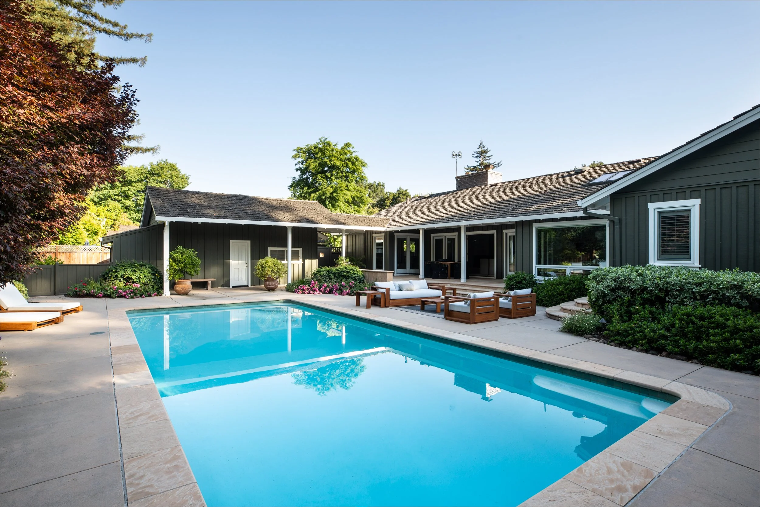 Backyard with in-ground swimming pool, patio seating area with chairs and cushions, lush greenery, and a house with dark siding and a covered patio