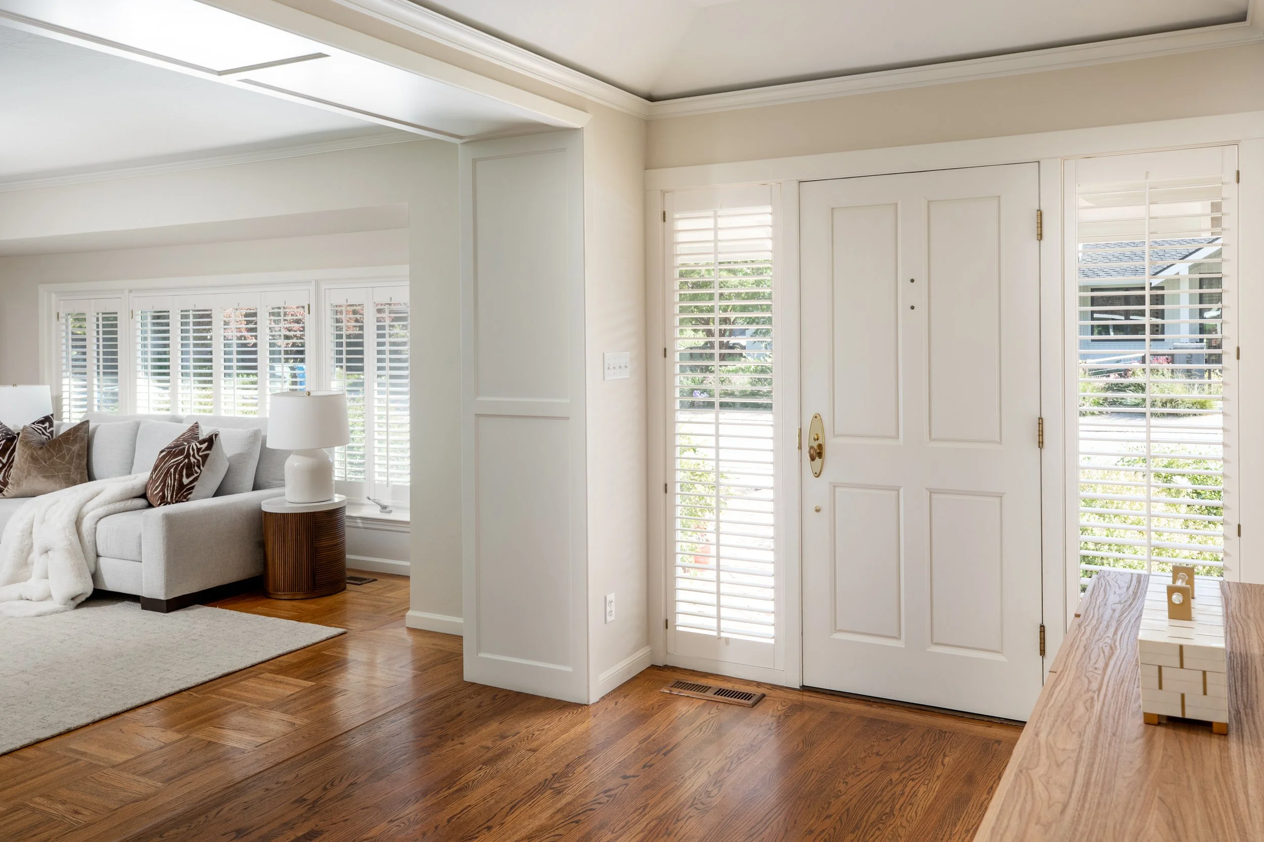 View of an entryway with white door and sidelights with plantation shutters, hardwood flooring, adjacent living room area with a sofa, pillows, and a lamp.