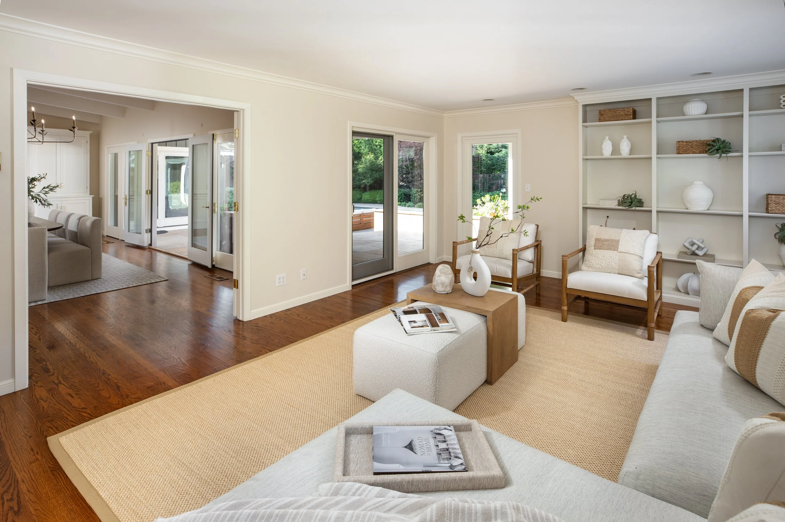 A living room with wooden floors, a beige sofa, a white armchair, and a coffee table with magazines. There's a large built-in shelf with decorative items and a sliding glass door leading to an outdoor patio with greenery.