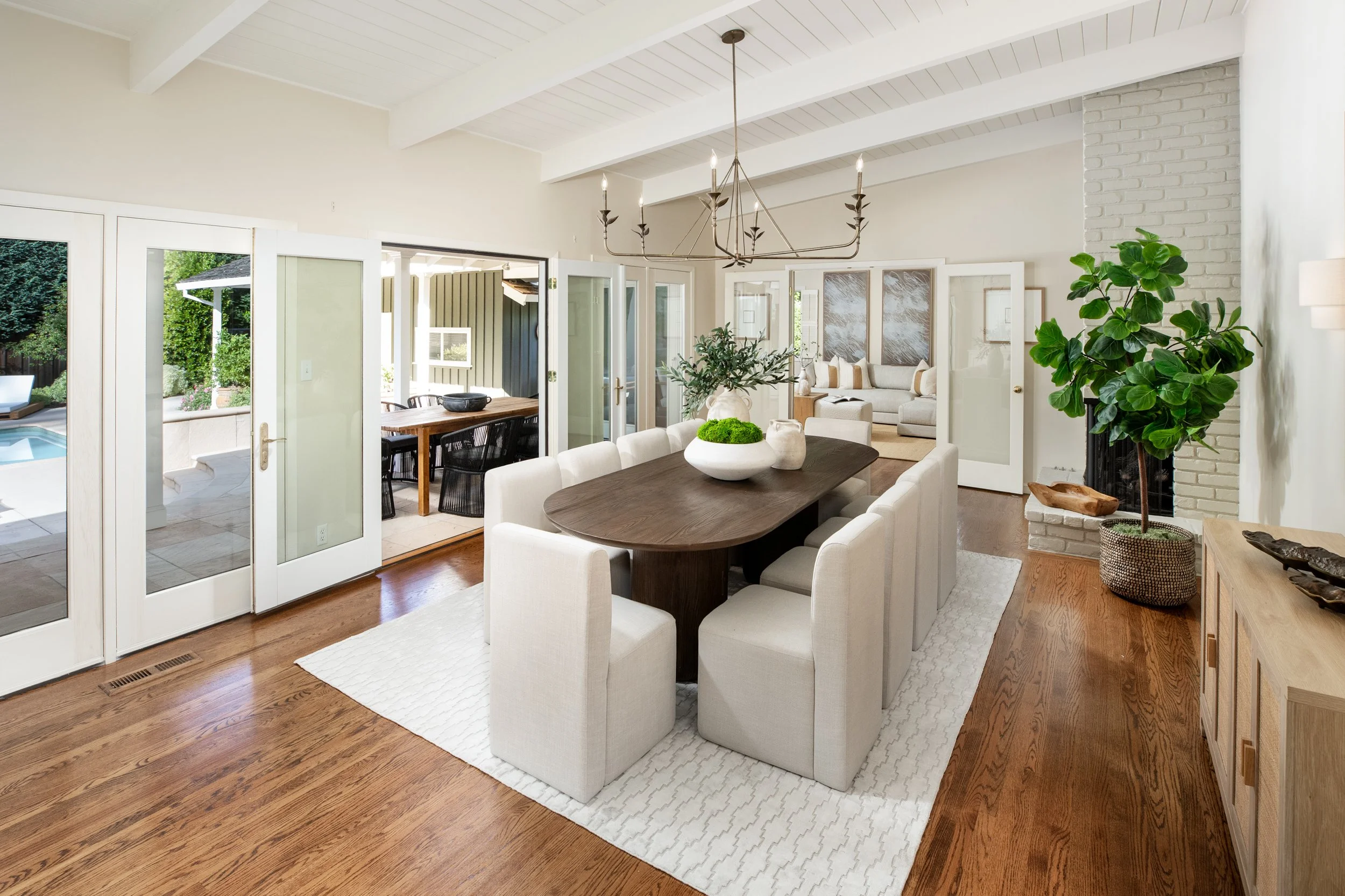 Bright living room with white walls, wooden floors, a dark wood dining table with white chairs, large glass doors opening to a patio with outdoor dining area and greenery, a chandelier hanging from the ceiling, and a tall potted plant near a white br