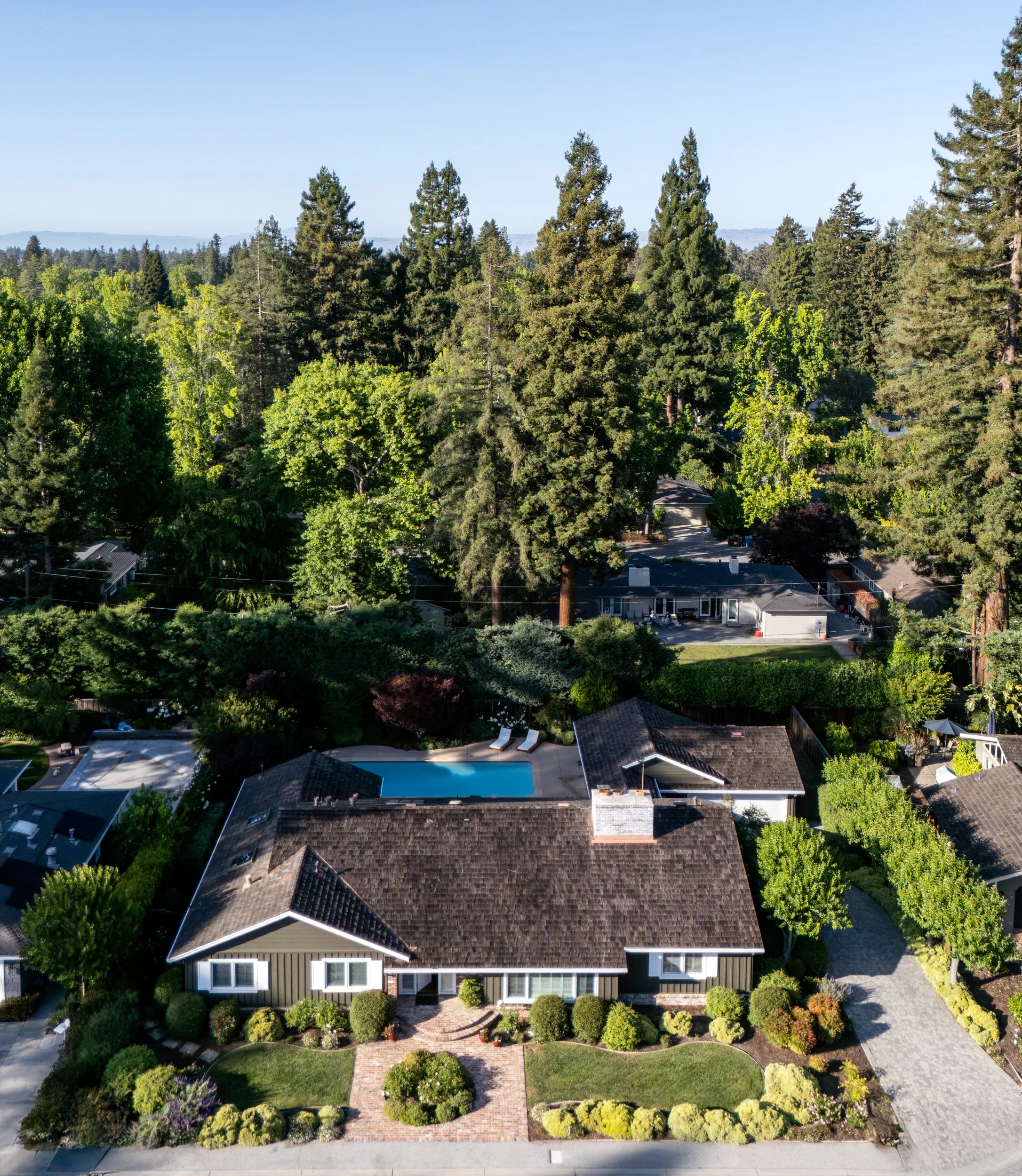 Aerial view of a residential neighborhood with multiple houses, lush green trees, and a swimming pool, under a clear blue sky.