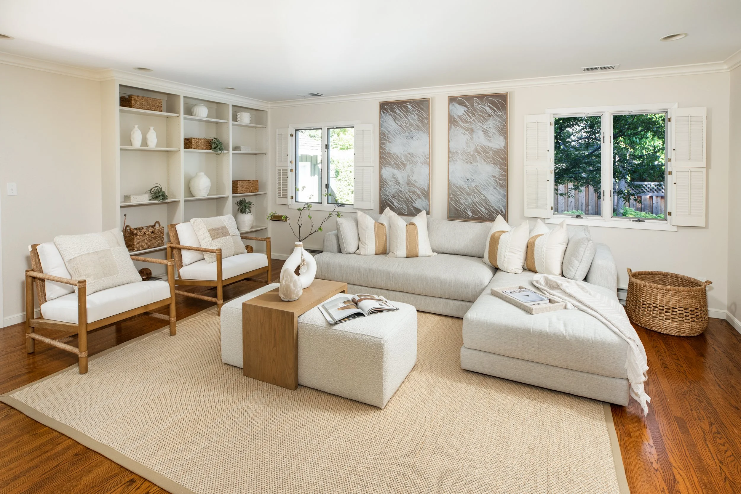 Living room with white and beige furniture, including a couch, chairs, and a coffee table, with decorative pillows, artworks on the wall, and large windows with white shutters showing a backyard. There are built-in shelves with white vases and basket