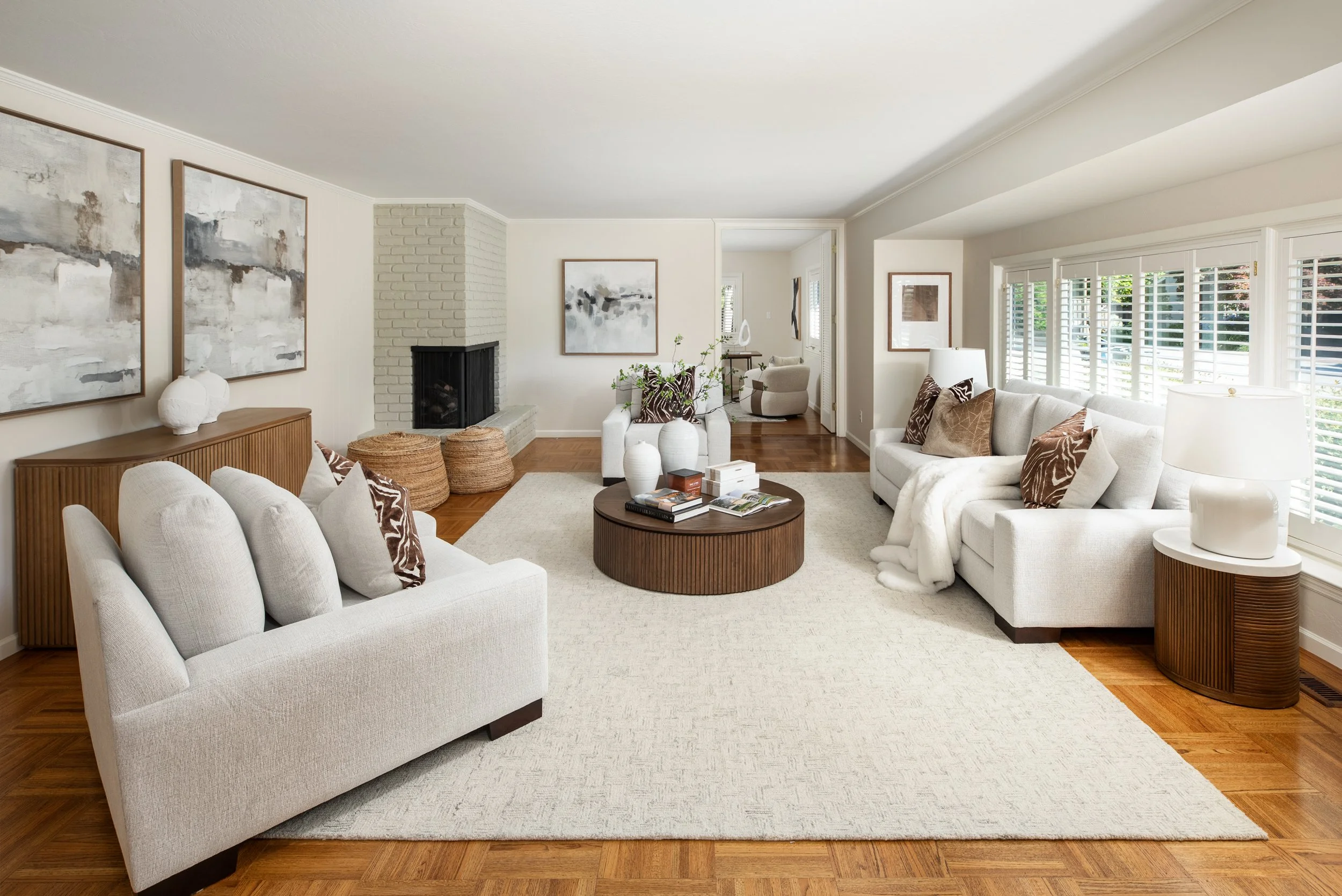 A living room with white sofa, armchair, wooden coffee table, abstract artwork, brick fireplace, and large windows with white plantation shutters.