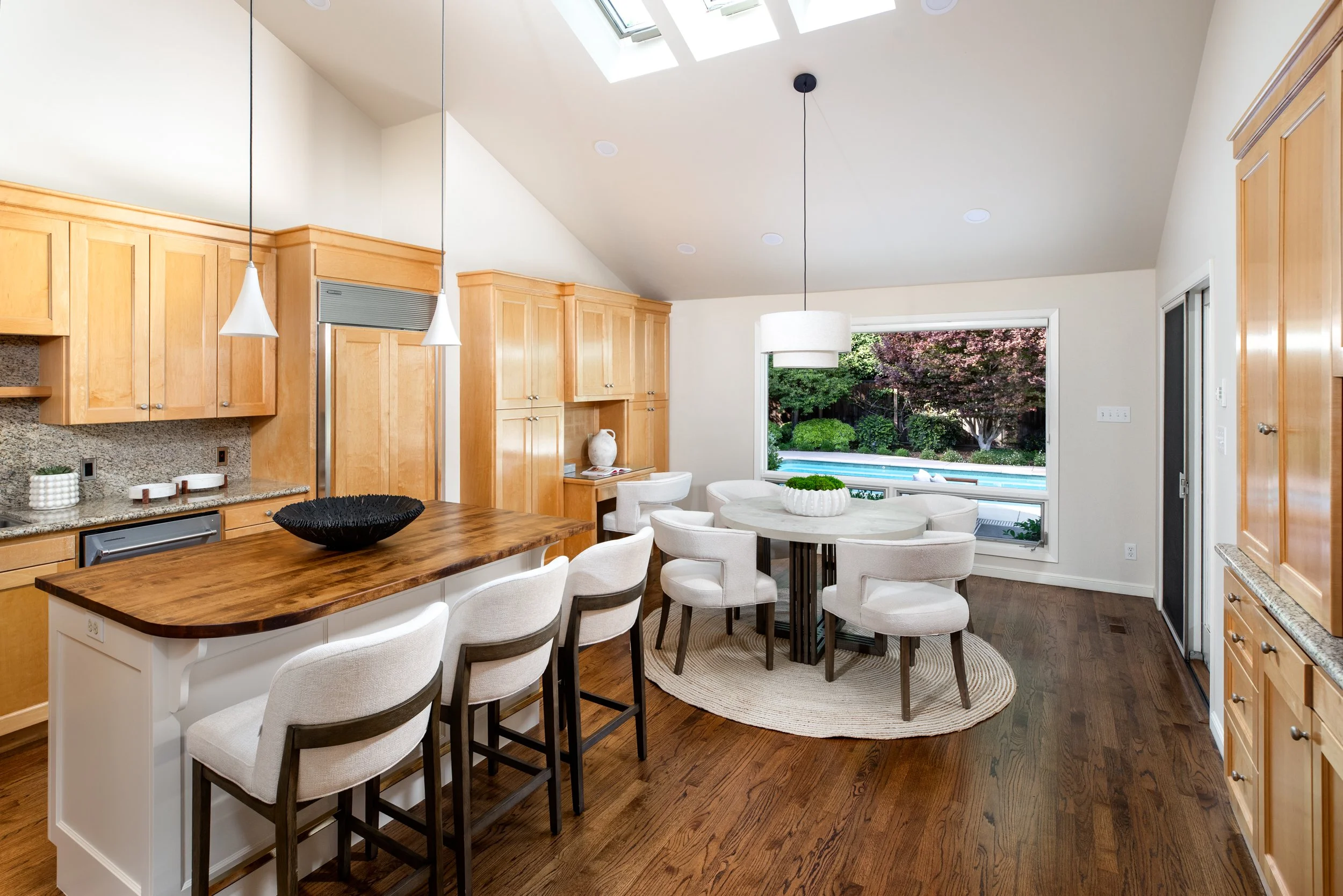 A bright kitchen and dining area with wooden cabinets, a granite countertop, a central island with bar stools, a round dining table surrounded by white upholstered chairs, and large windows showing a garden and swimming pool outside. Skylights are on