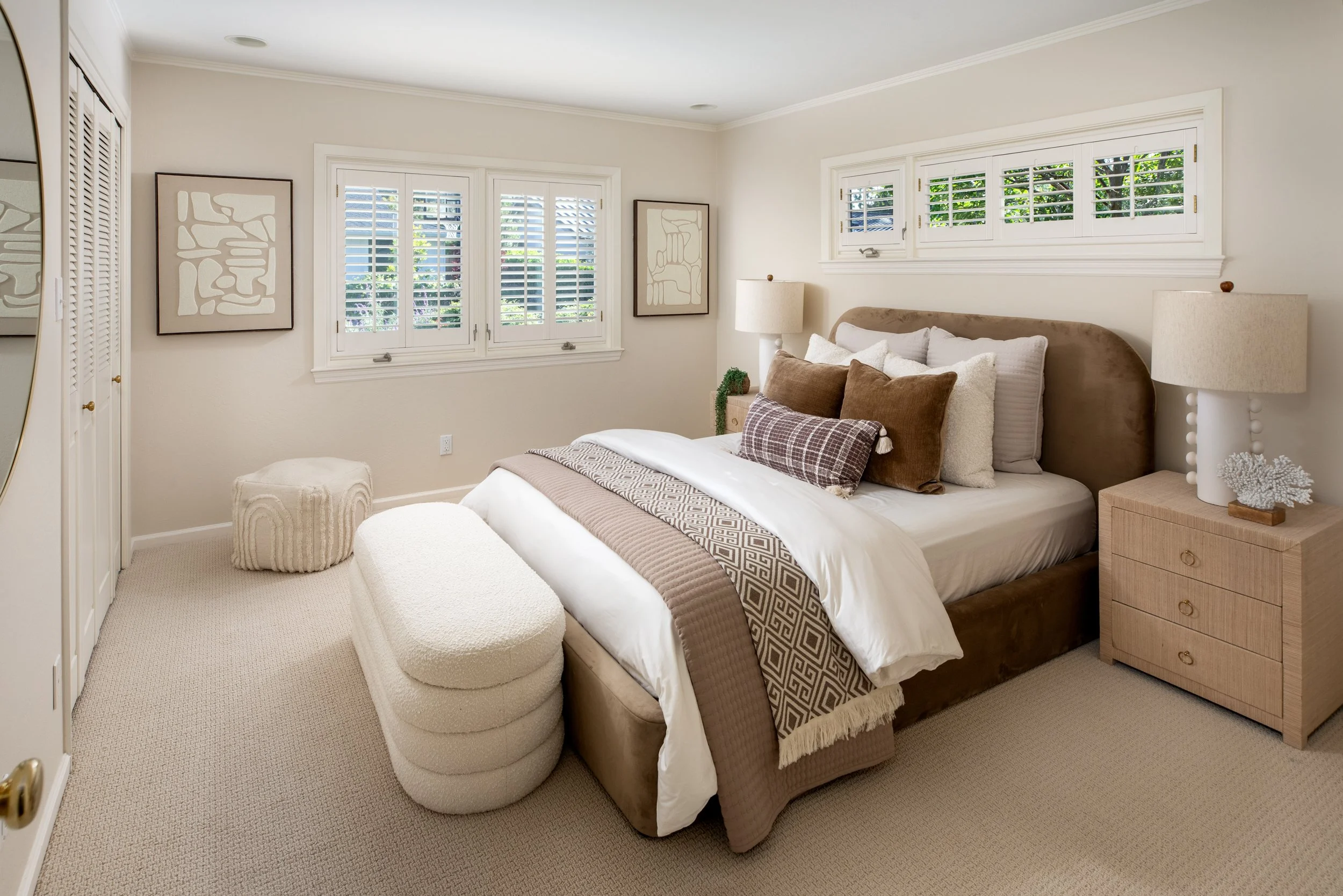 A cozy bedroom with a large bed, multiple pillows, two nightstands with lamps, abstract art on the walls, windows with white shutters, beige carpet, and a storage closet with louvered doors.