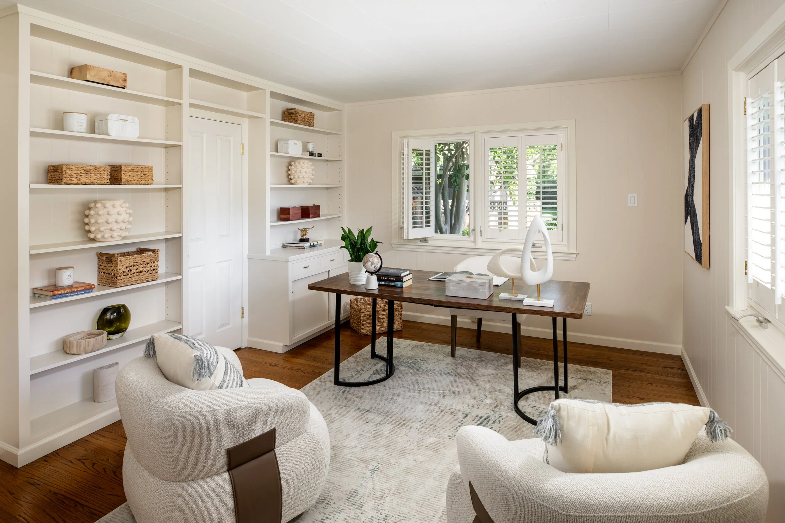A cozy living room with beige armchairs, a wooden table, white bookshelves filled with decorative items, and a window with white shutters overlooking green trees.