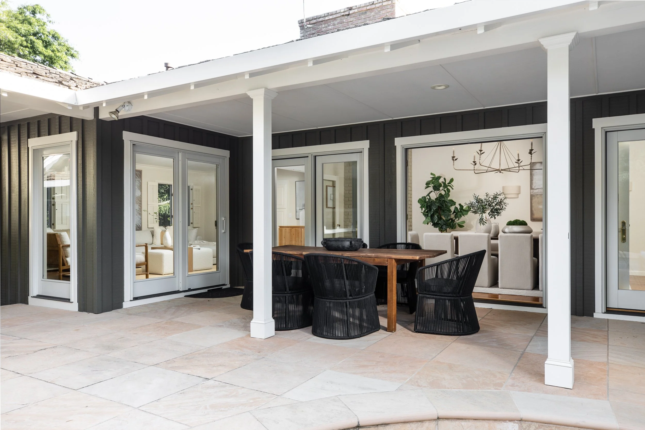 Outdoor patio with a dining table surrounded by black chairs, a view into a living room with plants, a chandelier, and modern decor.