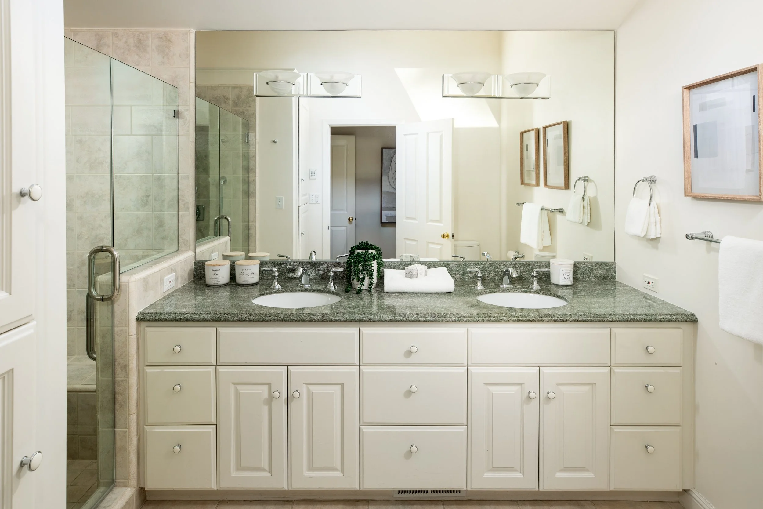 Bathroom with white vanity, granite countertop, double sinks, large mirror, towel bars, framed artwork, and a glass-enclosed shower.