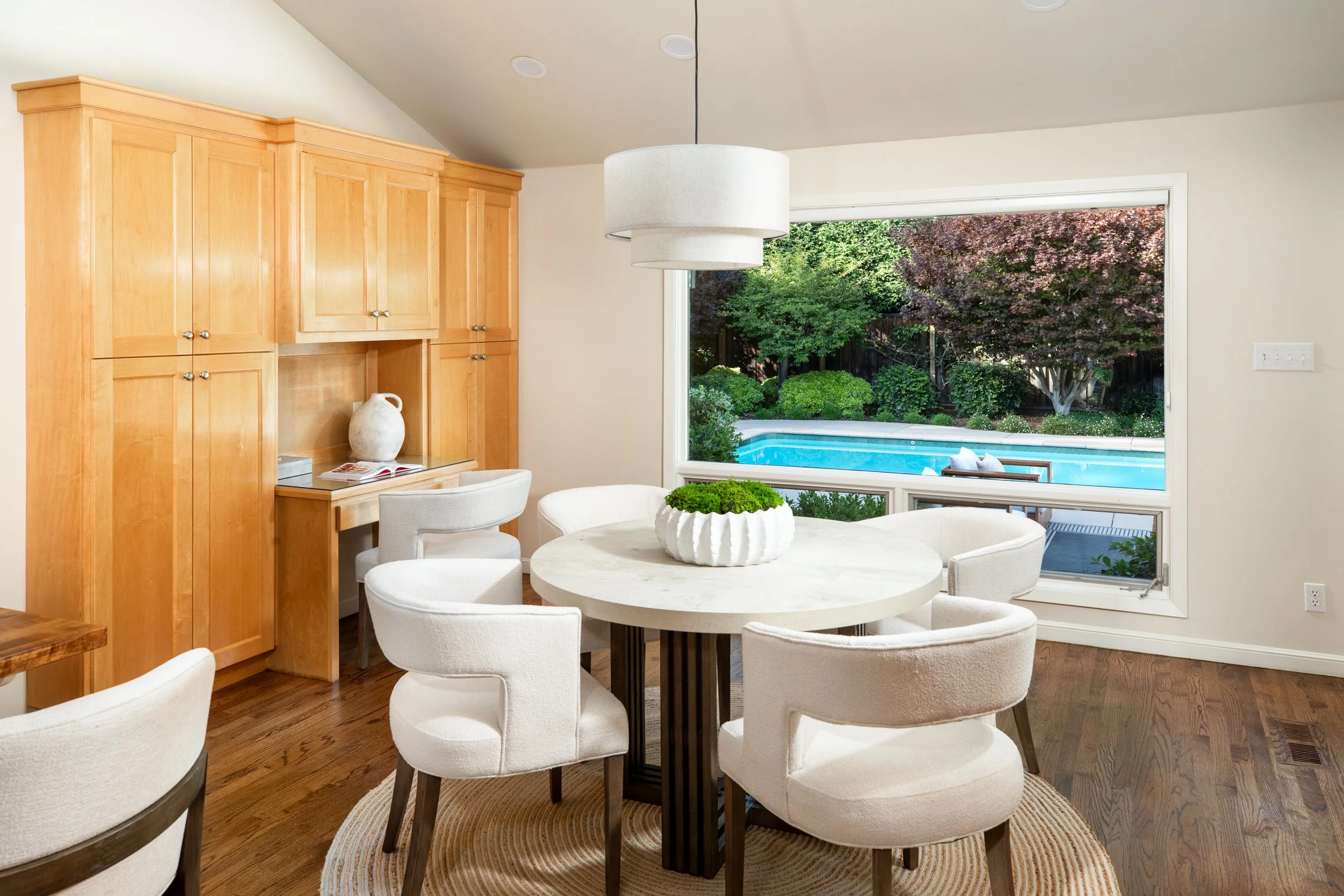 A dining area with a round table surrounded by white upholstered chairs, a large white ceramic vase on the table, a window showing a backyard with trees and a swimming pool, and wooden cabinets on the left side of the room.