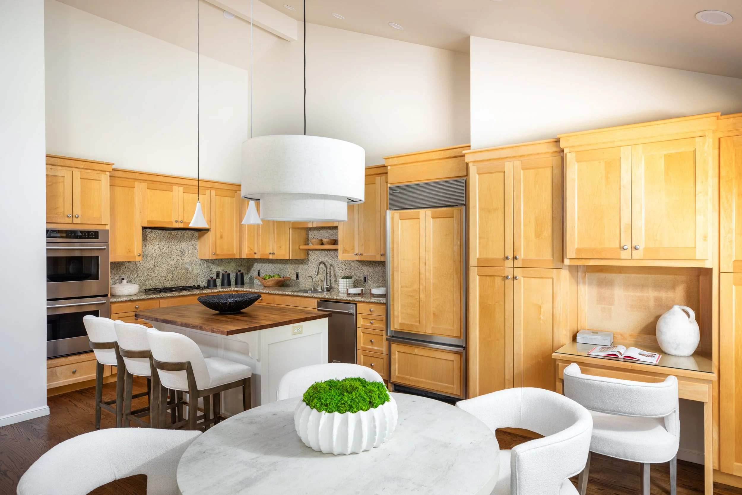 A kitchen with wooden cabinets, a white island with a wooden countertop, white chairs around a table with a white vase filled with moss, and modern appliances including double ovens and a built-in microwave.
