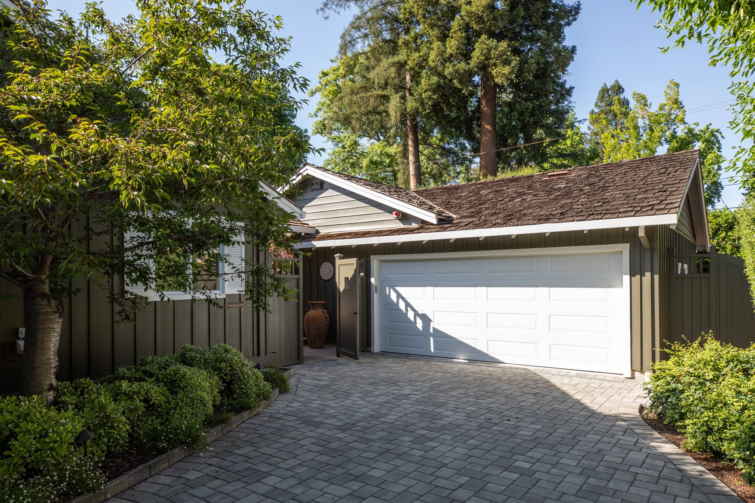 A residential garage with a white door, surrounded by a tree and green shrubs, under a clear blue sky.