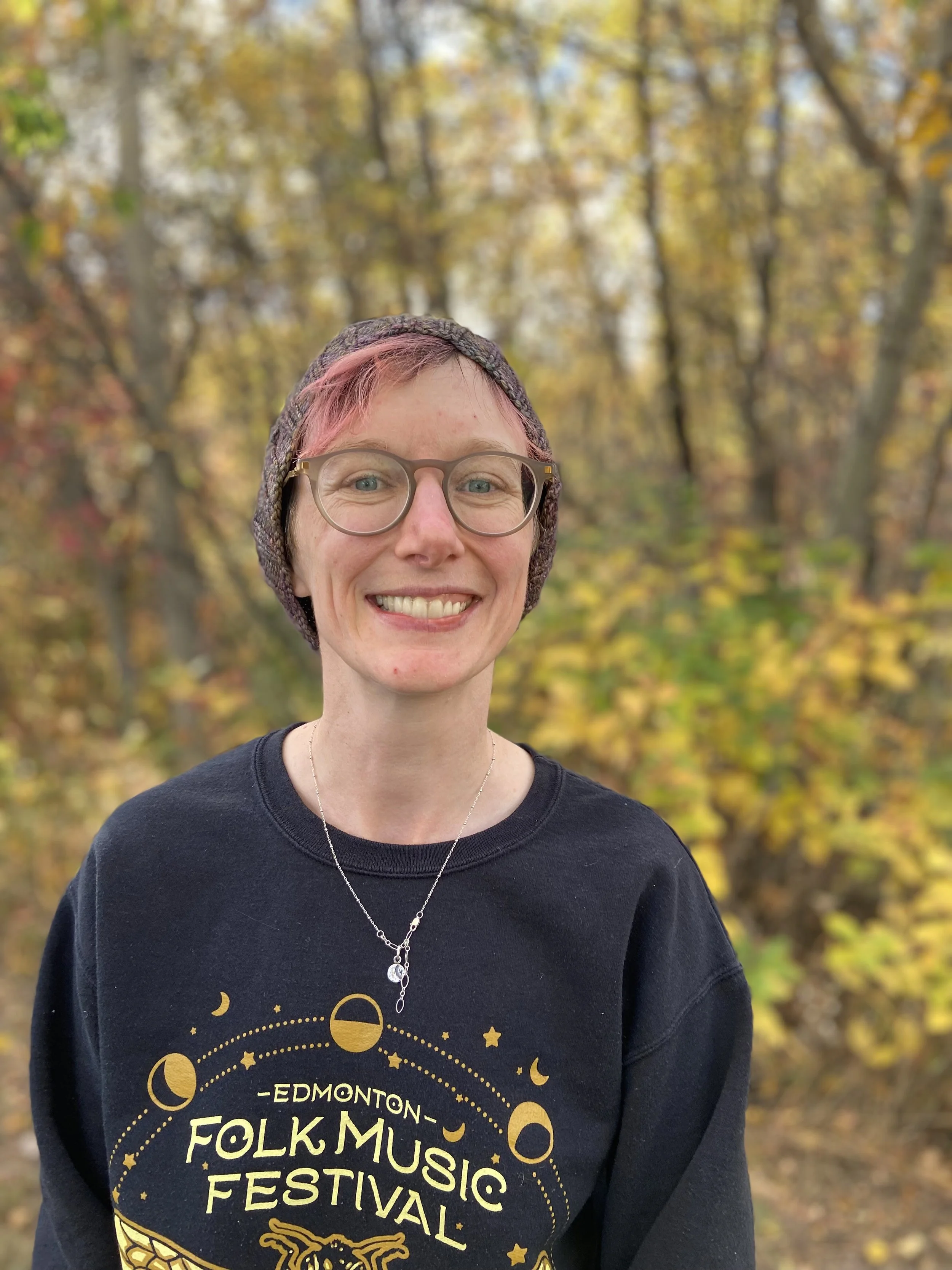 A person with short pink hair, glasses, and a knit toque smiling outdoors. They are  wearing a black sweatshirt with yellow lettering and a silver necklace. The background shows trees with autumn yellow and orange leaves.
