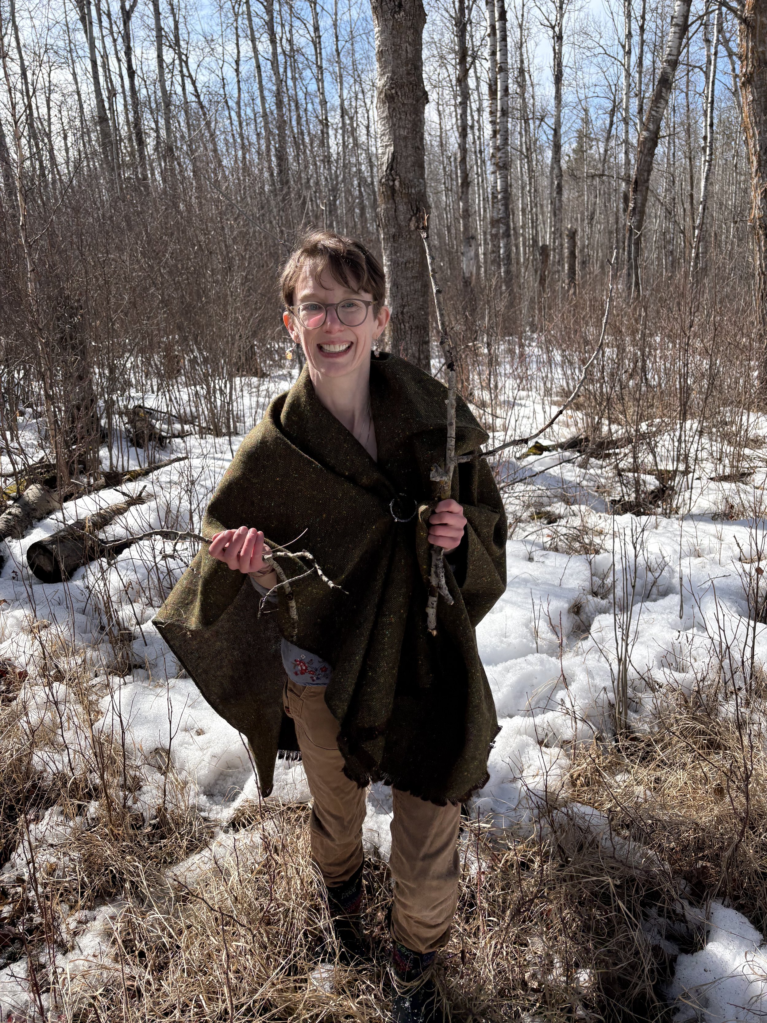 Person  with short brown hair, glasses, and earrings standing outdoors in a snowy forest. They are smiling, holding a small branch, and wearing a dark green cloak and khaki pants.