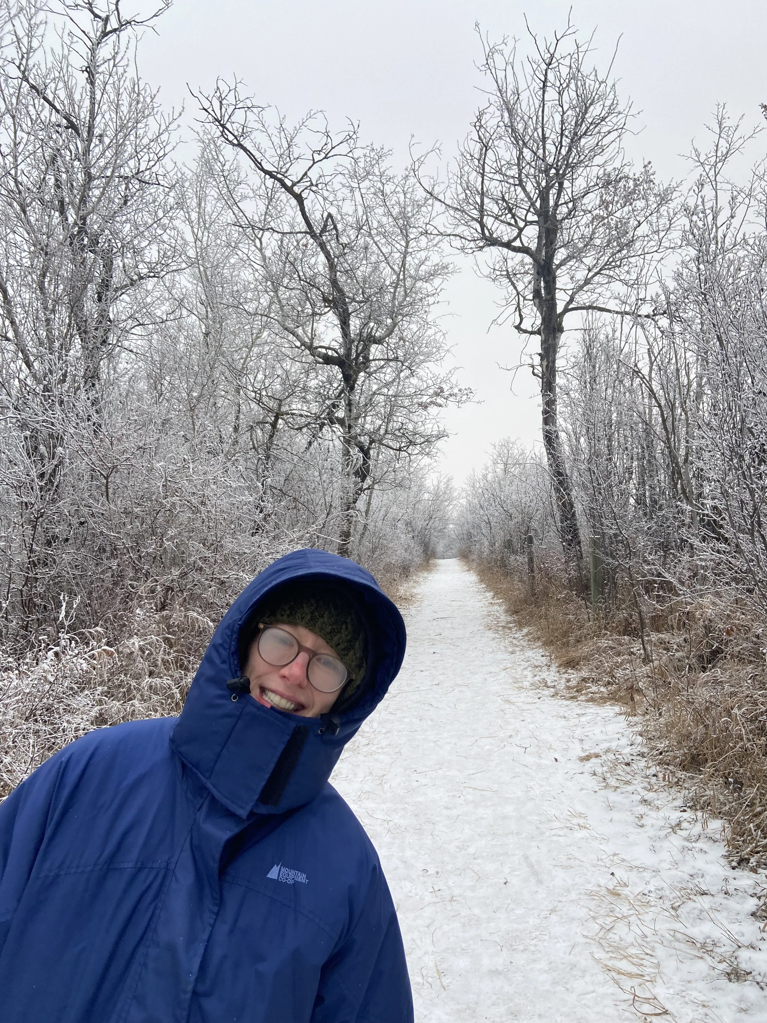A person wearing glasses, a knit hat, and a blue hooded snow  jacket, smiling outdoors on a snowy winter trail surrounded by snow-covered trees.