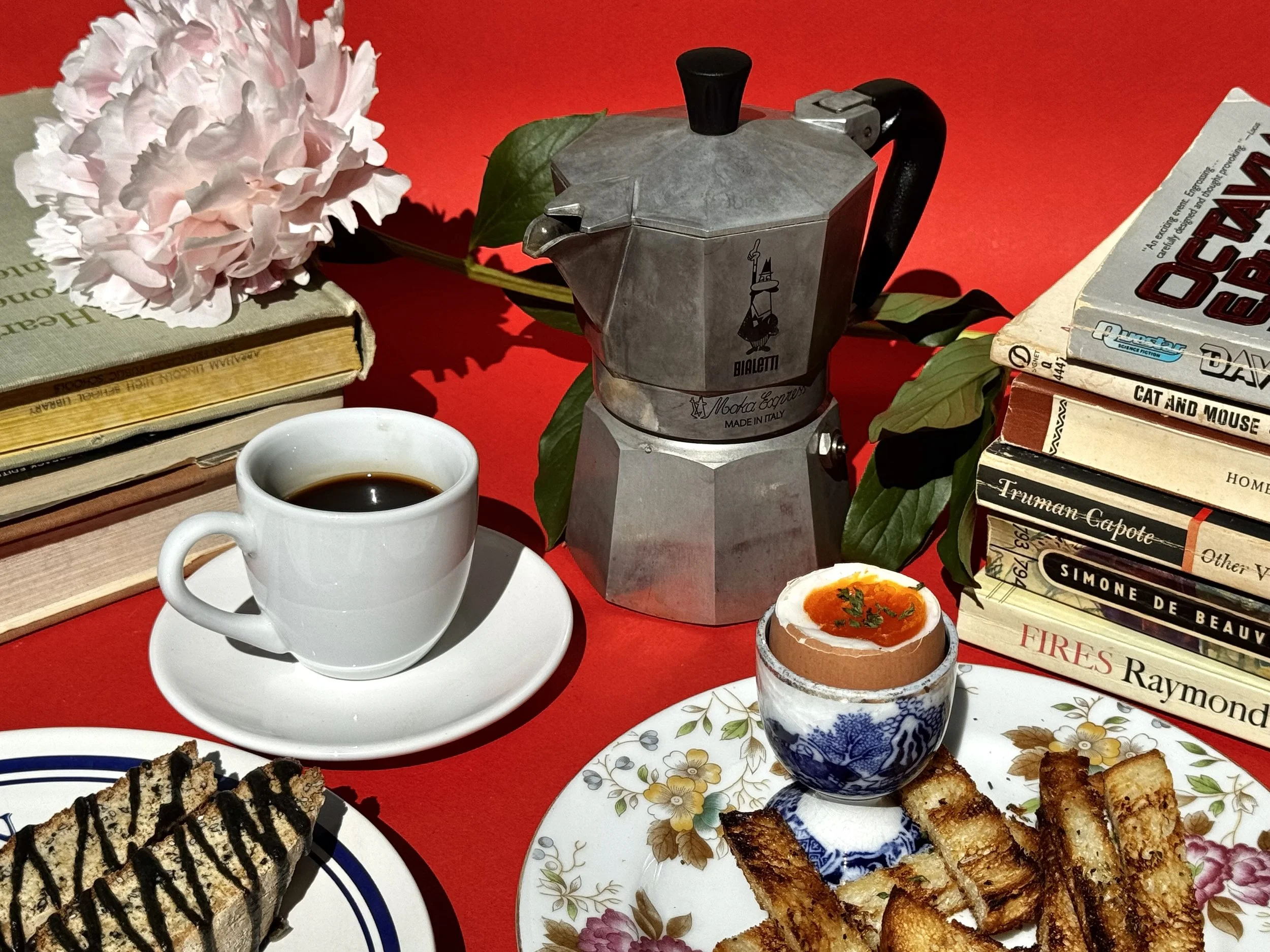Stacks of books, a pink peony flower, a moka espresso coffee maker, a cup of black coffee, a soft boiled egg plate, and a plate of biscotti on a red background.