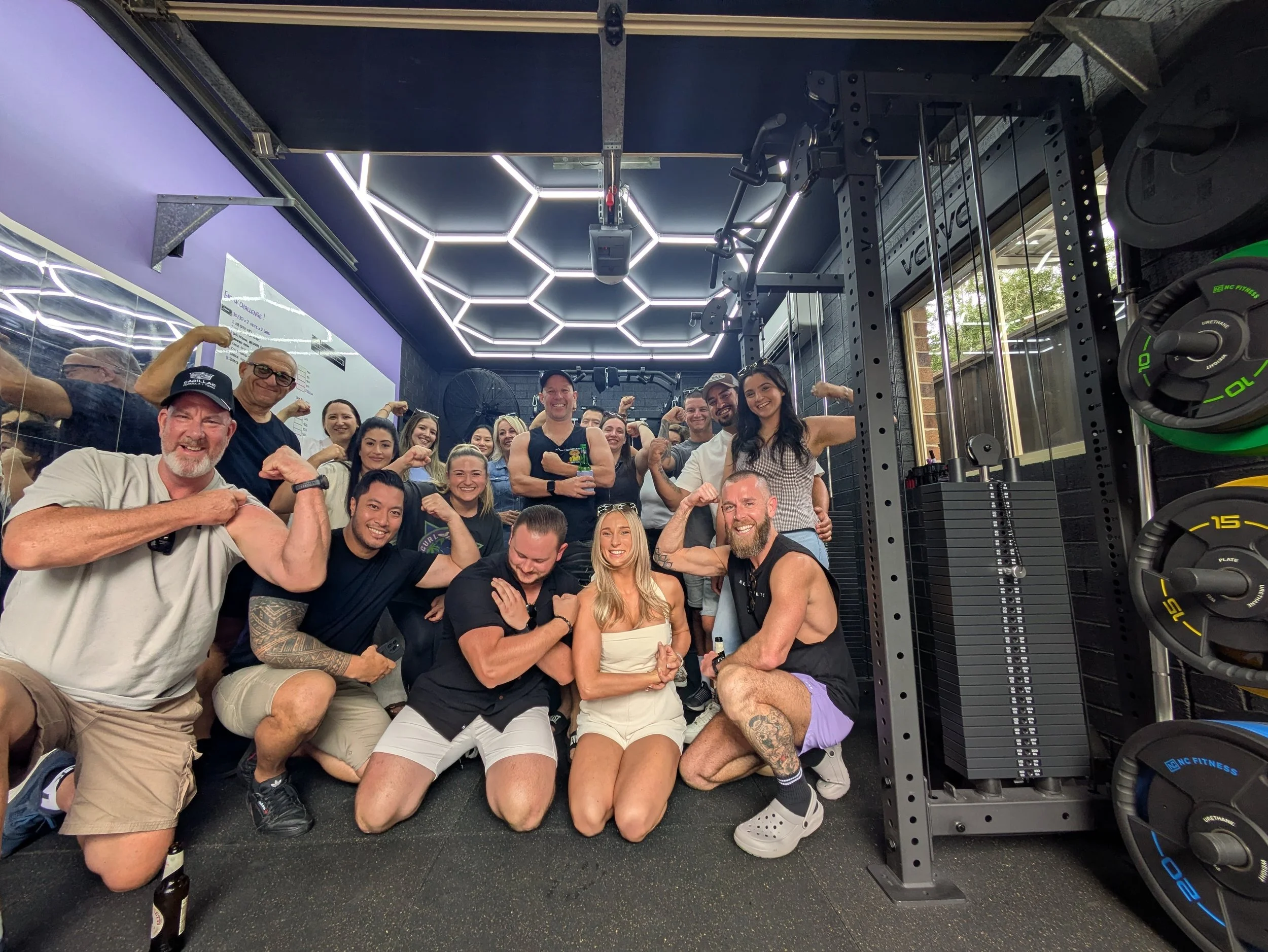 A group of diverse people posing in a gym with flexed muscles, surrounded by workout equipment, with a ceiling decorated with illuminated hexagon patterns.