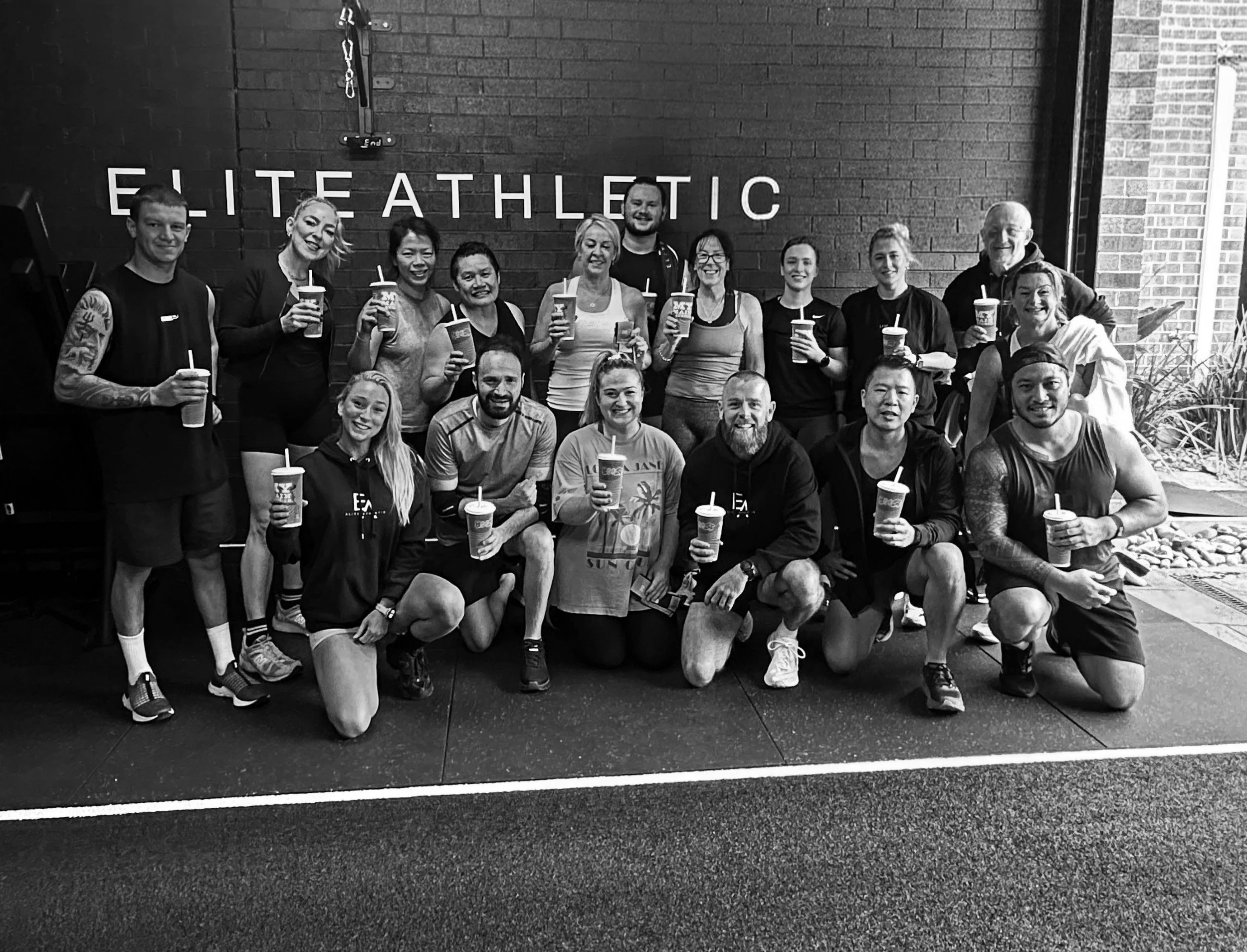 A group of 16 people in athletic clothing, holding drinks with straws, posing for a photo inside a gym with a brick wall that has the words 'ELITE ATHLETIC' on it. Some people are kneeling in front, others are standing behind.