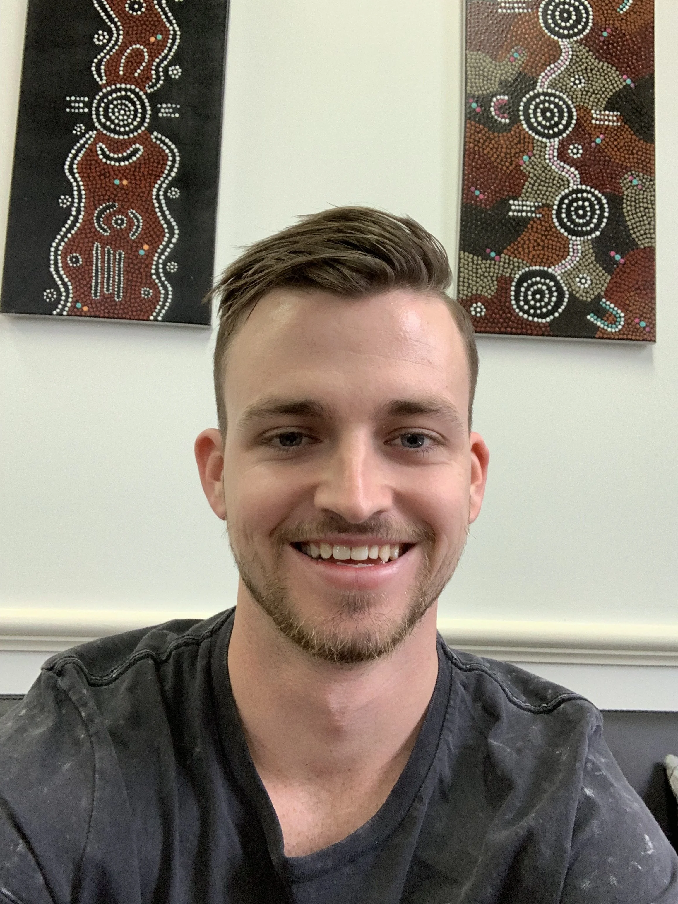 COSupports Director, Brandon Dunn, with short brown hair, beard, and mustache, wearing a black shirt. Behind him are two Aboriginal-style dot paintings on the wall.