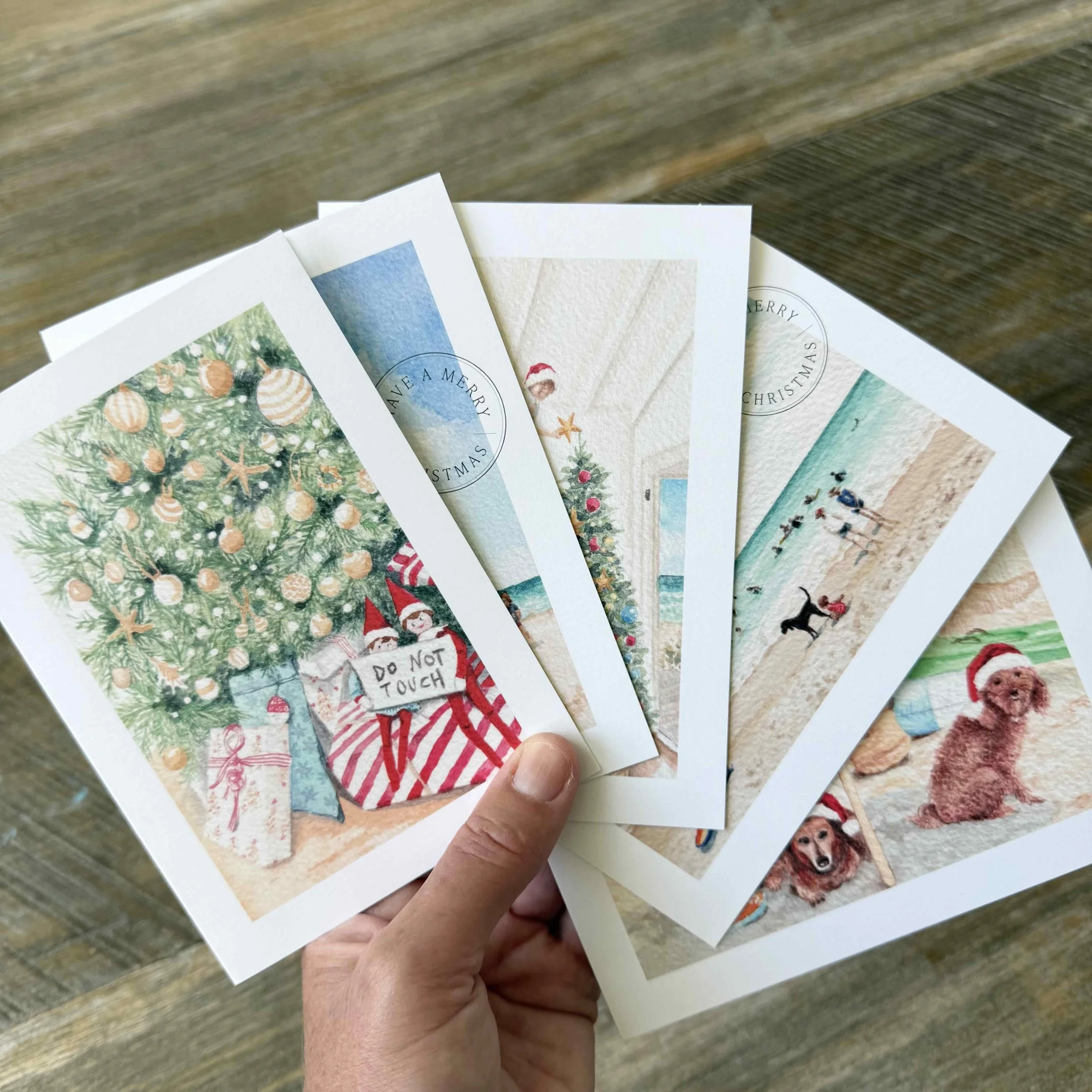 Five original Salt Brush Coastal Australian Christmas card designs displayed in a hand, showing the premium printed watercolour greeting cards.