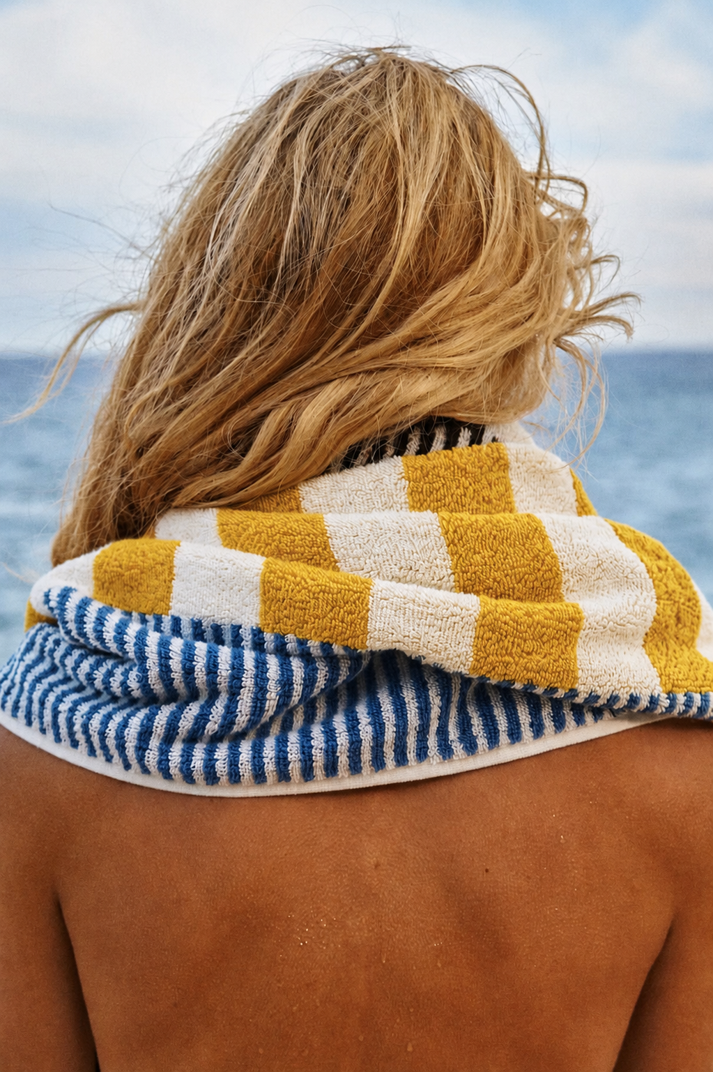 A woman with blonde, windswept hair standing outdoors near the ocean, wrapped in a yellow and white checkered towel over her shoulders, with a partial view of her back and shoulder.