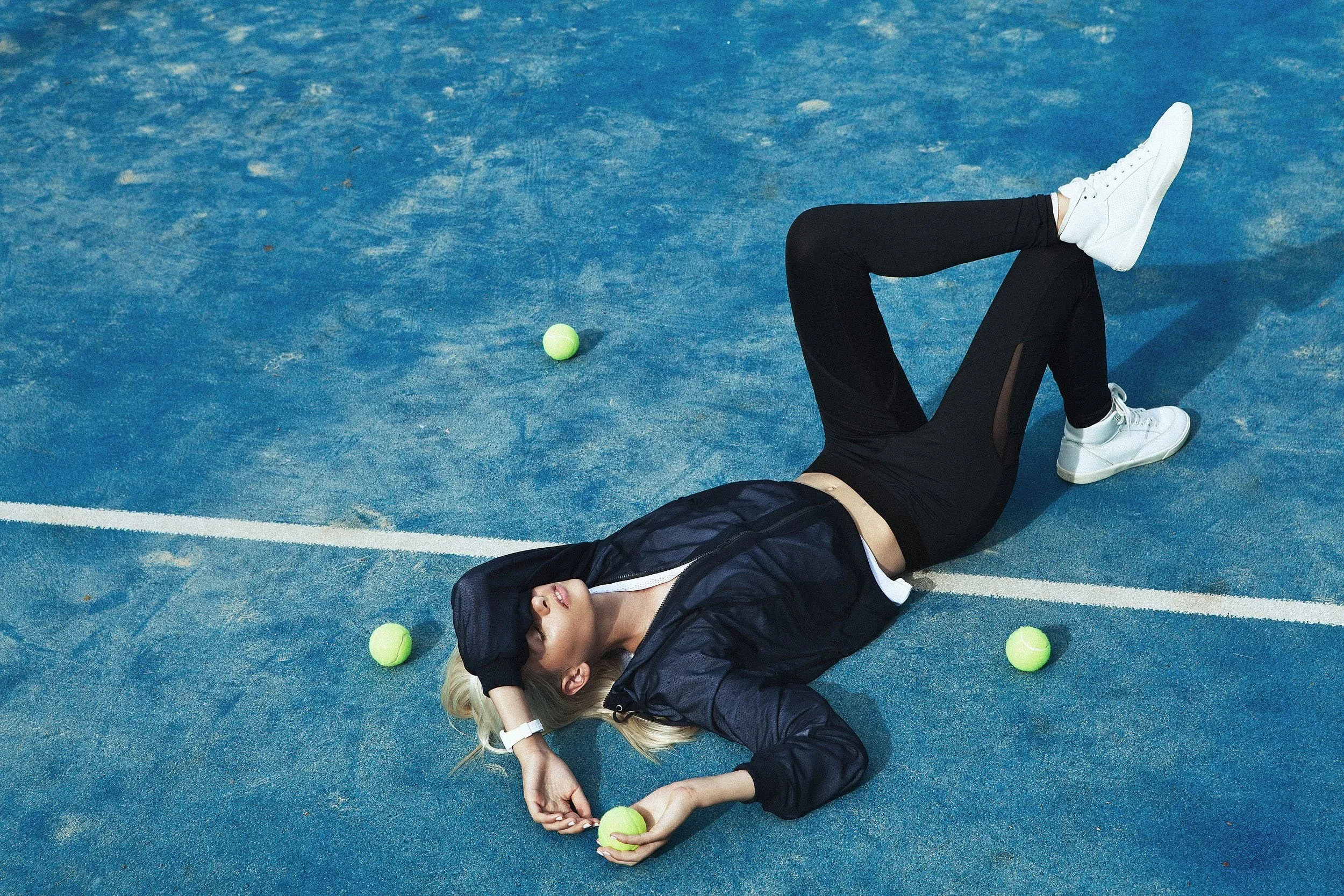 A young woman lying on her back on a blue tennis court with three tennis balls around her, wearing a black jacket, black pants, and white sneakers.