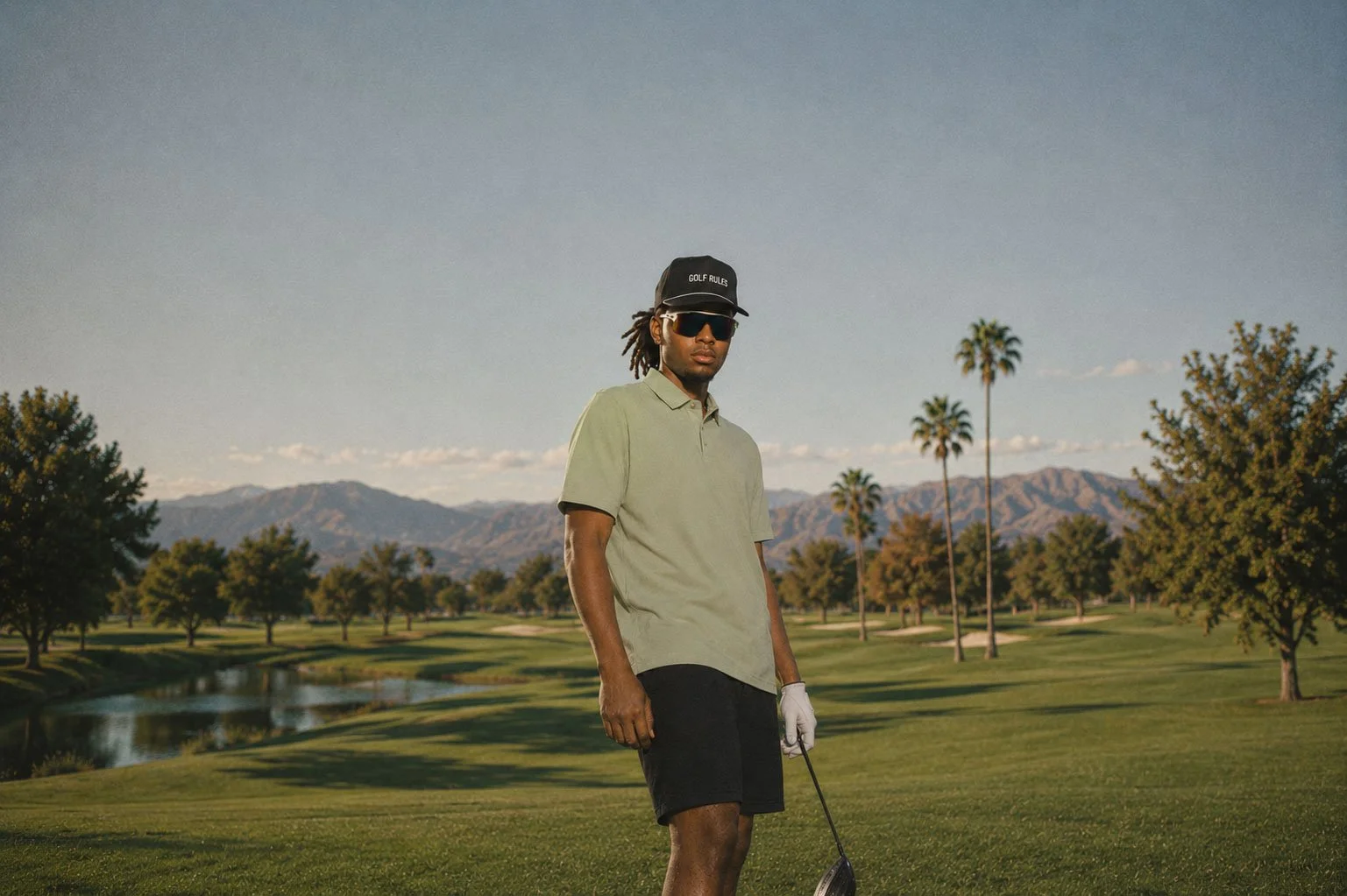 A man with sunglasses, a black cap, and a golf glove holding a golf club on a golf course with palm trees, mountains, and a lake in the background.