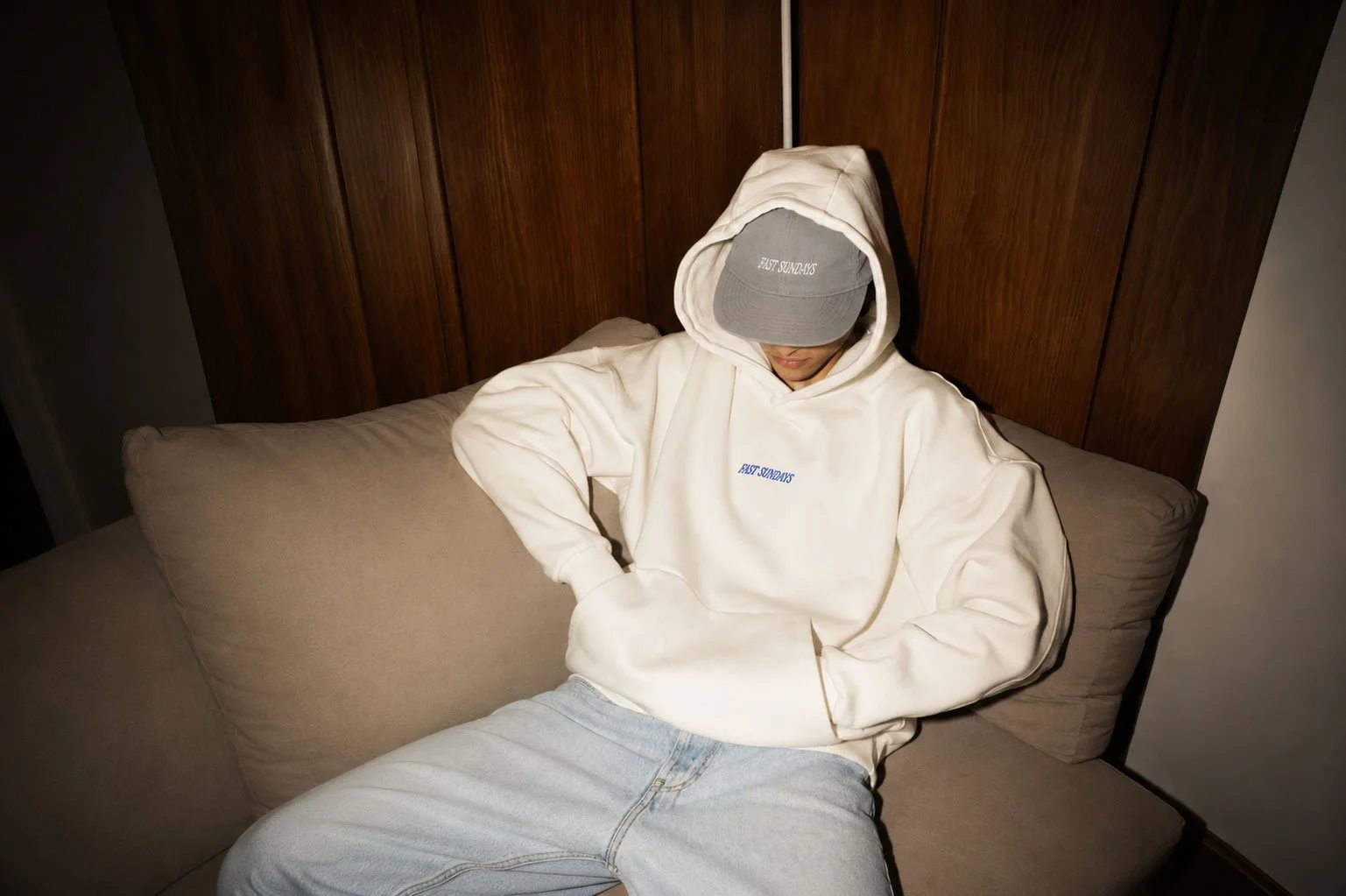 Person sitting on a beige sofa against a wooden wall, wearing a white hoodie with the hood up, a gray cap with white lettering, and light blue jeans, with their head tilted downward.