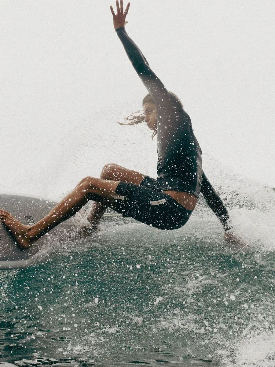 A person in a black wetsuit is captured mid-fall off a surfboard on a wave, with water splashing and their hand raised in the air.