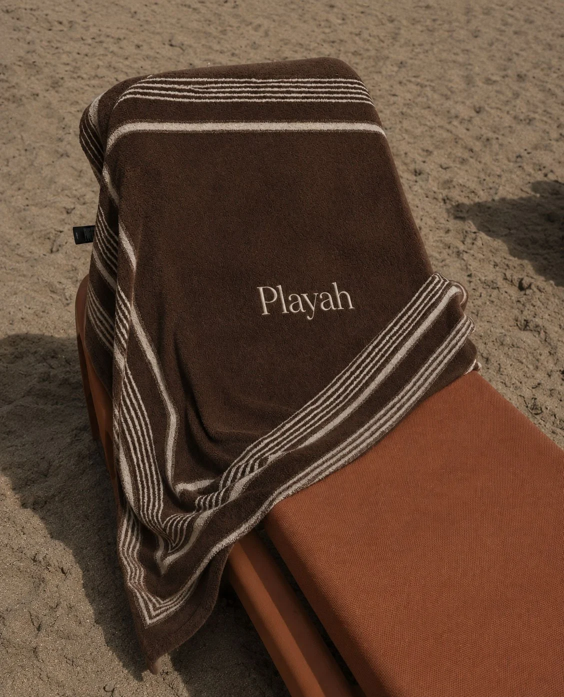 A brown lounge chair on sandy ground with a dark brown towel draped over it, embroidered with the name 'Playah' in white.