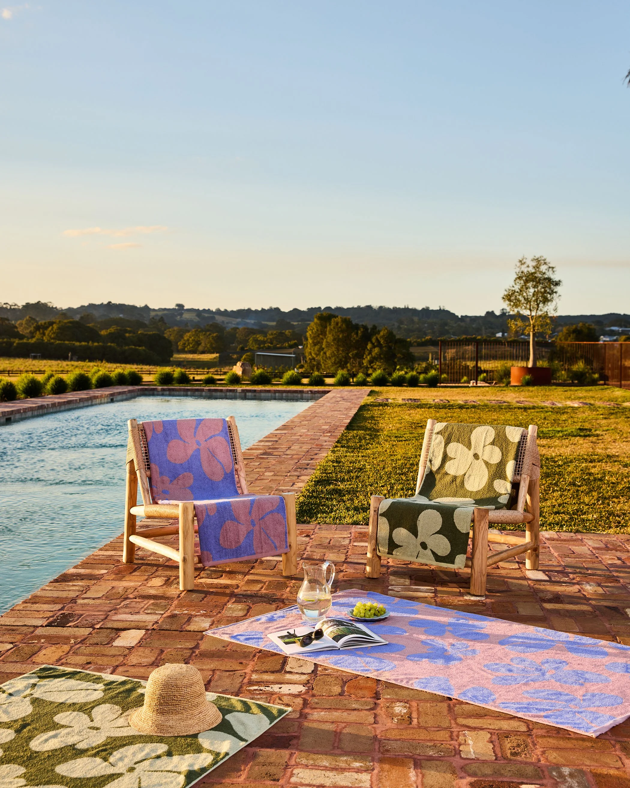 Outdoor scene featuring two chairs with floral-patterned towels, a water pitcher, a book, and sunglasses on a brick patio near a pool. In the background, there are rolling hills, trees, and a clear sky.