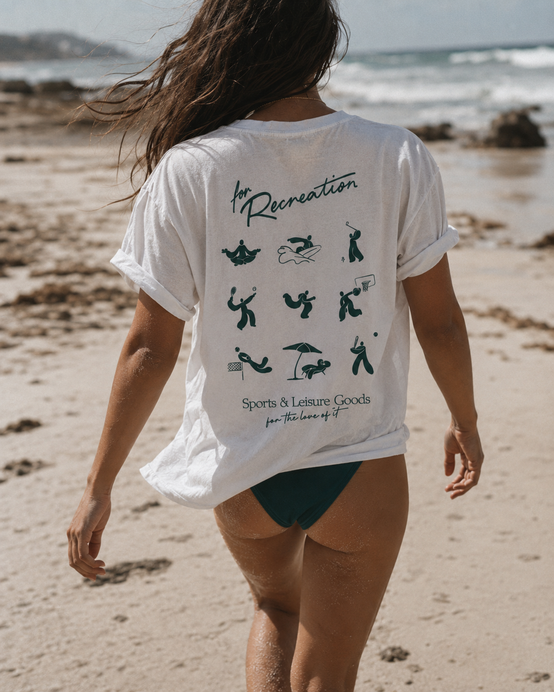 Person walking on beach wearing a white T-shirt with sports and leisure icons on the back.