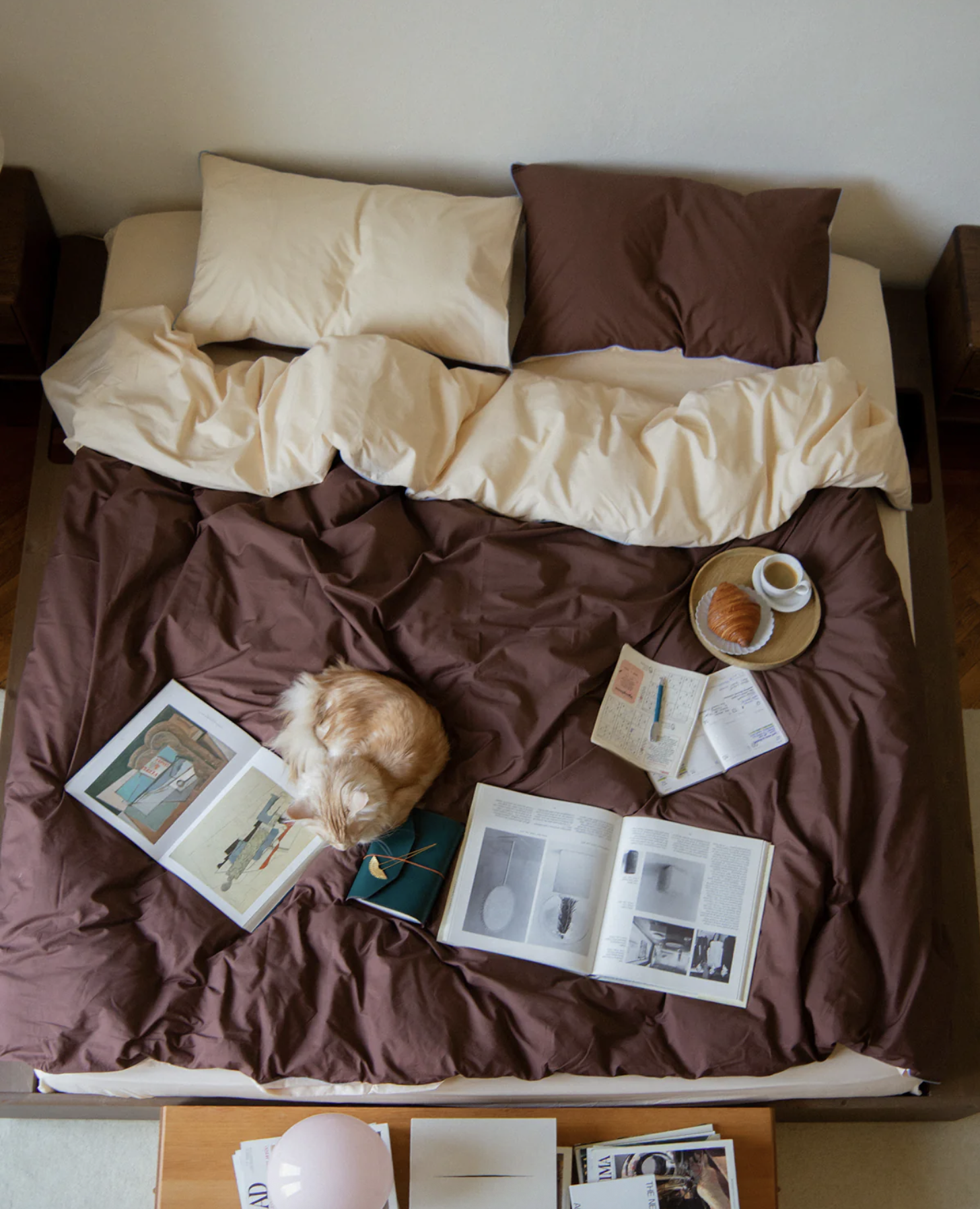 A bed with beige and brown bedding, two pillows, and an open magazine. A cat is curled up on top of the magazine. There are books and papers on a small wooden table at the foot of the bed, with a pink spherical lamp on it. A tray with a croissant, a cup of coffee, and a plate is on the bed. The wall behind is plain and light-colored.