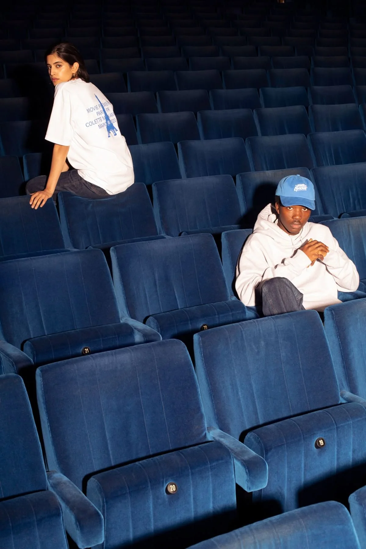 Inside an empty theater with blue velvet seats, a young woman with dark hair and a white T-shirt sitting on a seat looking over her shoulder, and a young man in a white hoodie and blue cap sitting alone, looking at the camera.