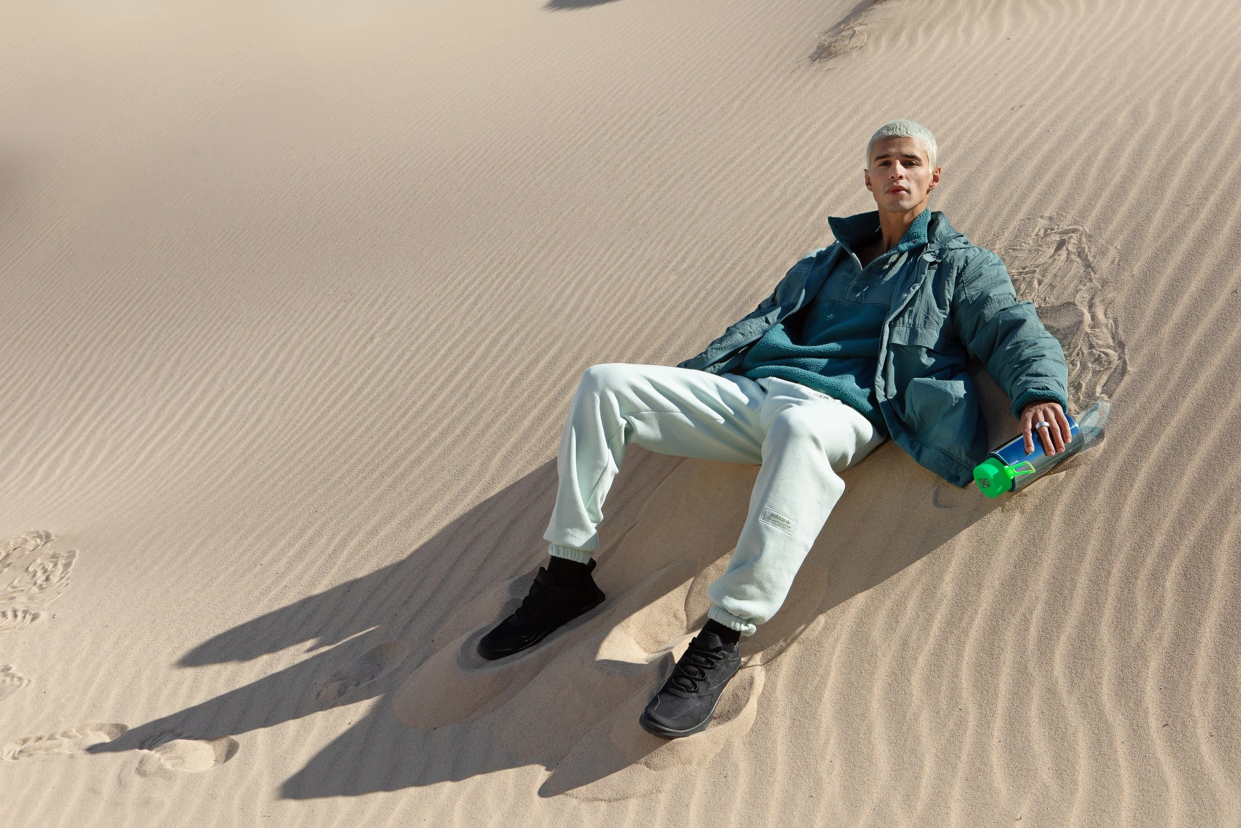 Man sitting on sand dune in a desert, wearing a blue jacket, white pants, black shoes, holding a green water bottle.