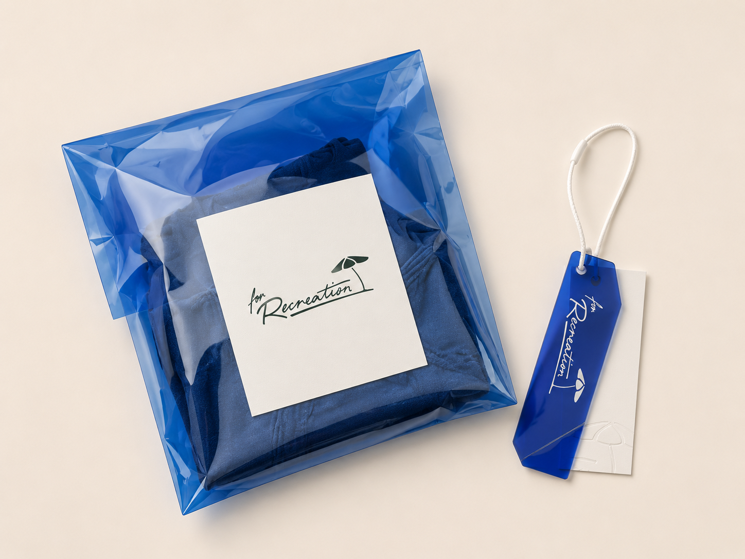 A neatly folded piece of clothing inside a clear blue plastic gift bag with a white card that reads 'for Recreation' and a blue tag with the same message attached to the bag.