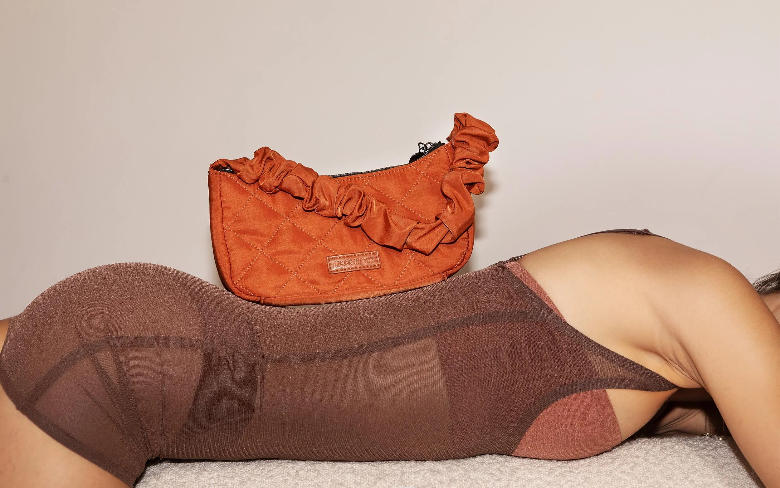 Close-up of a person lying face down on a beige carpet, wearing sheer, dark-colored tights, with a small orange quilted handbag with a ruched strap and black zipper on their back.