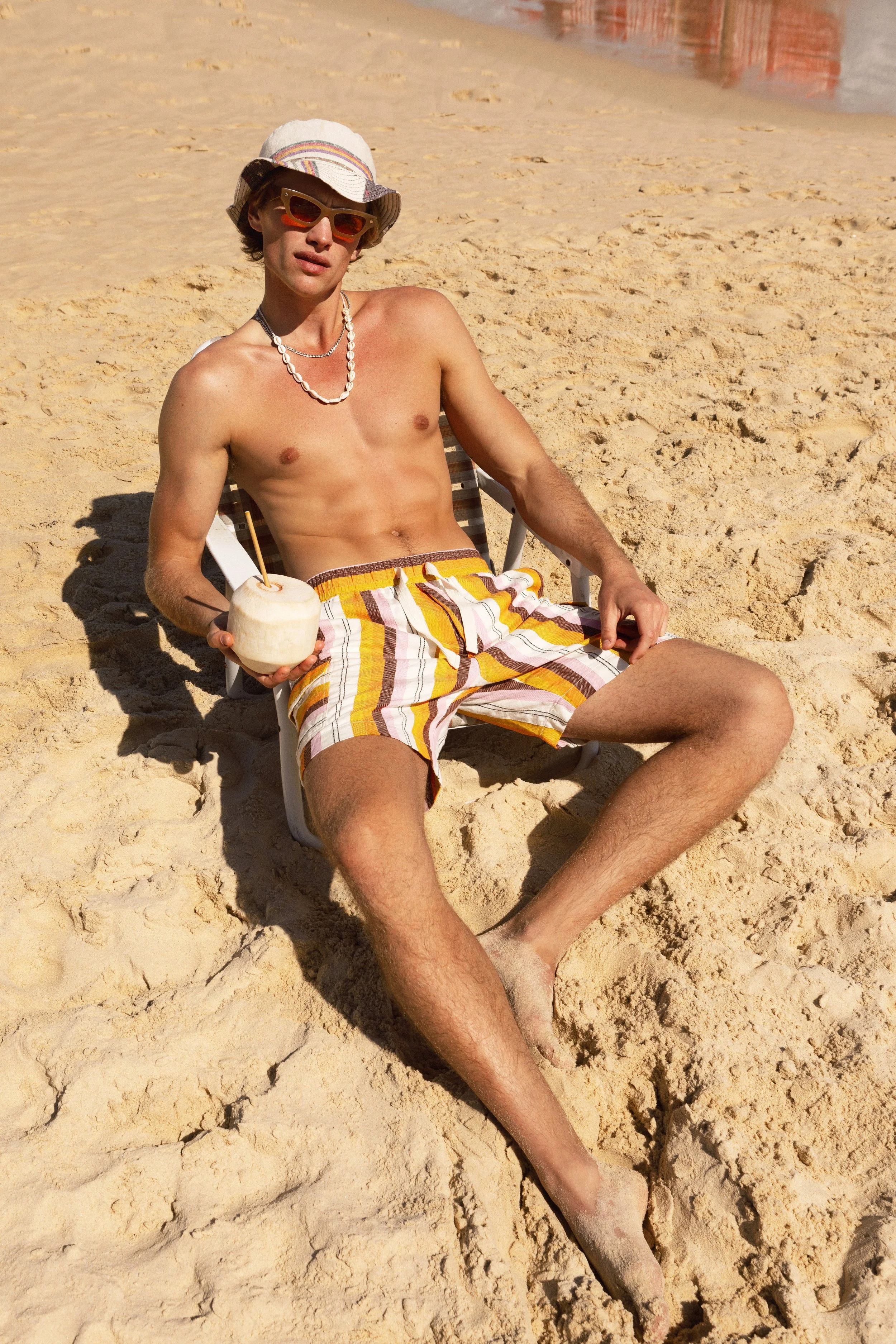 A young man relaxing on a beach chair on sandy beach, wearing striped shorts, a bucket hat, colorful sunglasses, and necklaces, holding a coconut with a straw.