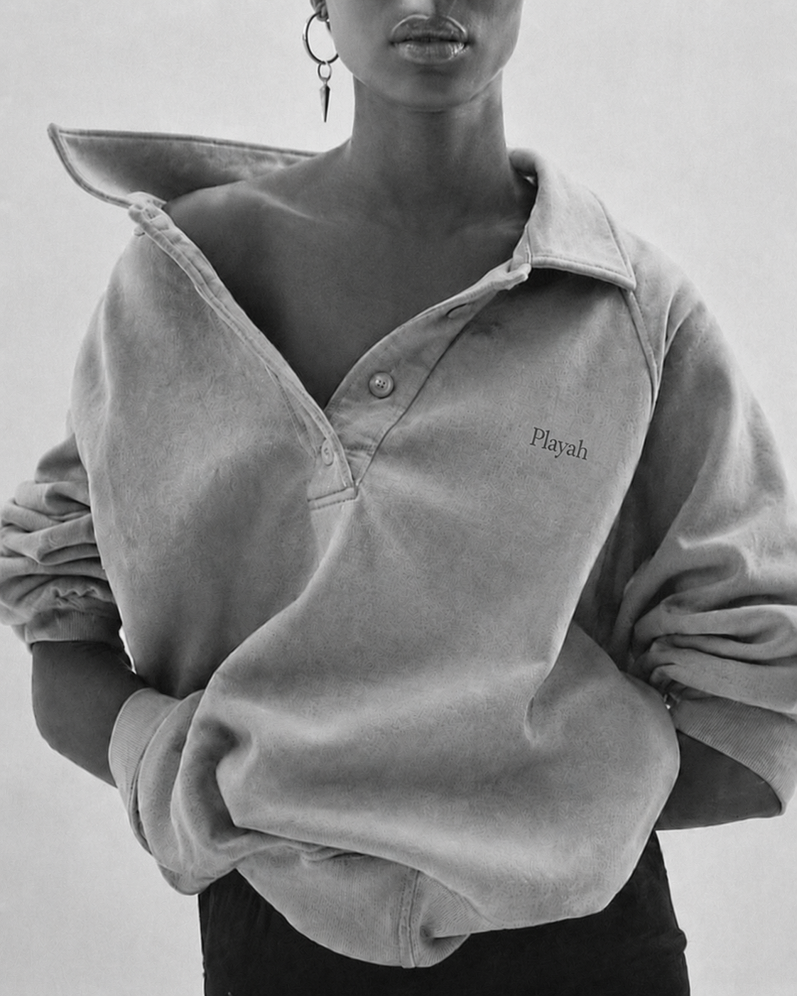 Black and white photo of a woman wearing a sweatshirt that has the name 'Playah' embroidered on it. One shoulder is exposed with a watermelon slice placed on it, and she is wearing an earring with a spike charm.