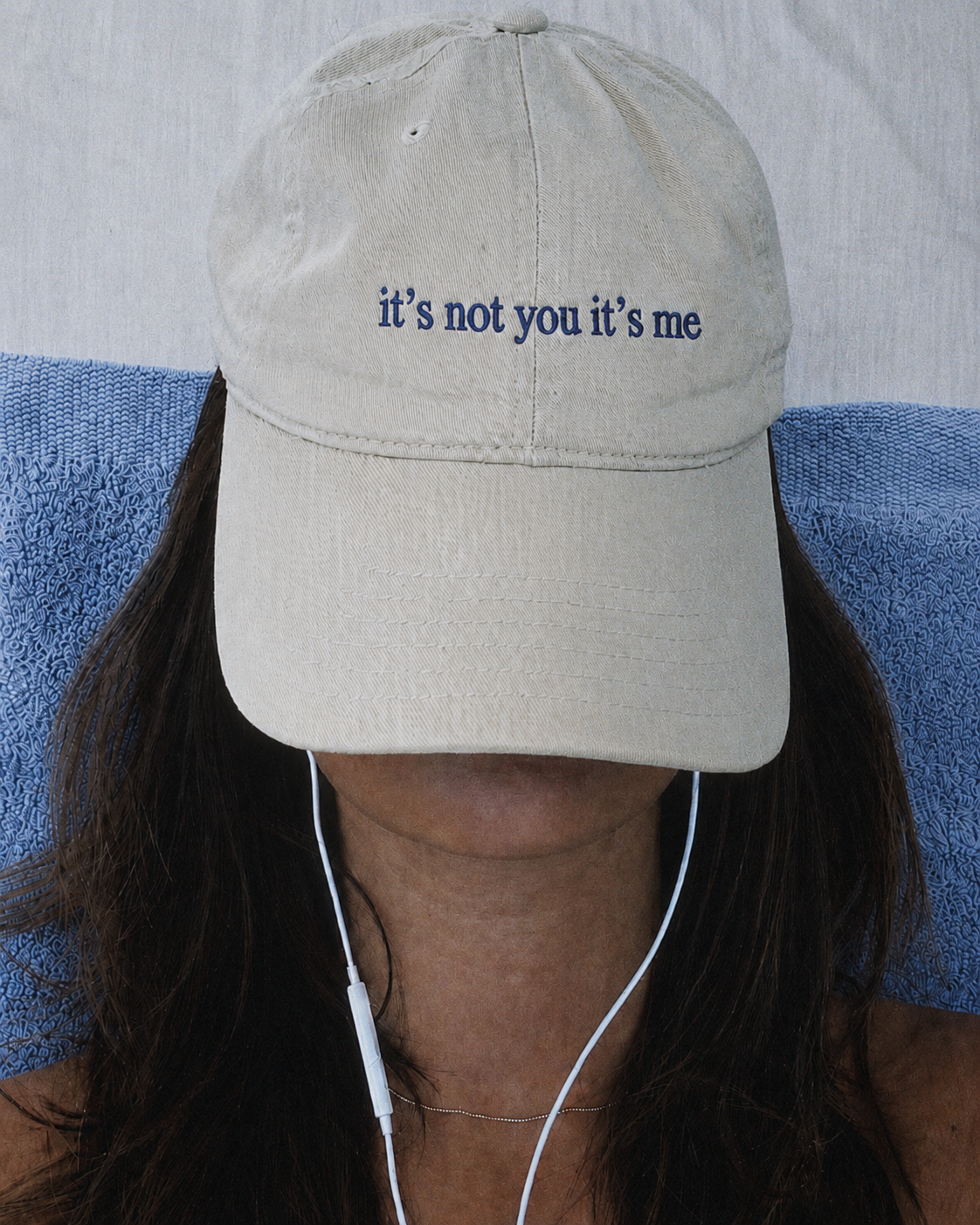 Person wearing a beige cap with the text "it's not you it's me" embroidered on it, sitting in a blue patterned chair, with dark hair, wearing white earphones.