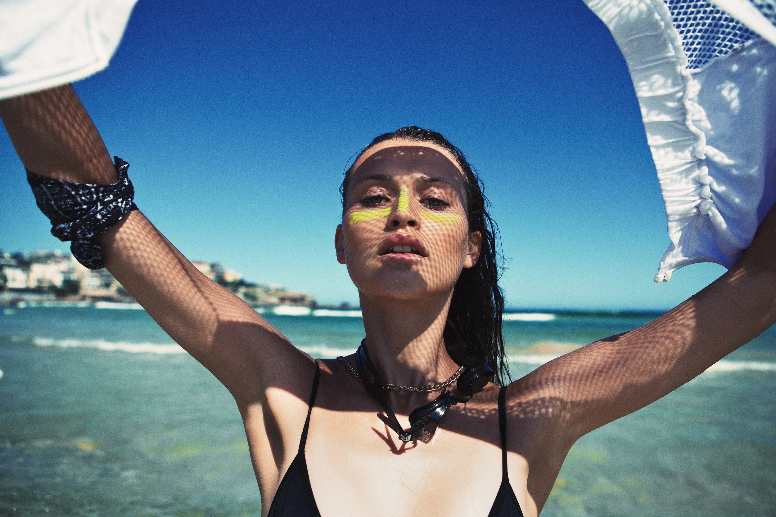 A woman with dark hair and face paint, standing on a beach with a blue sky, holding a white shirt or towel overhead, casting a shadow with a grid pattern on her face and arms.