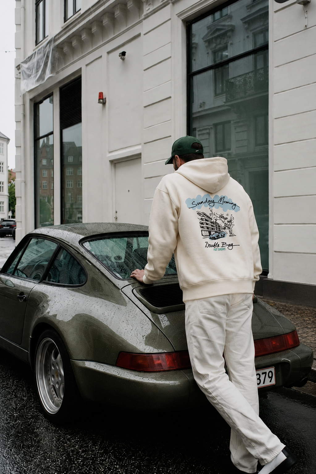 Man wearing a beige hoodie and matching pants cleaning a classic green Porsche car parked on a city street on a rainy day.