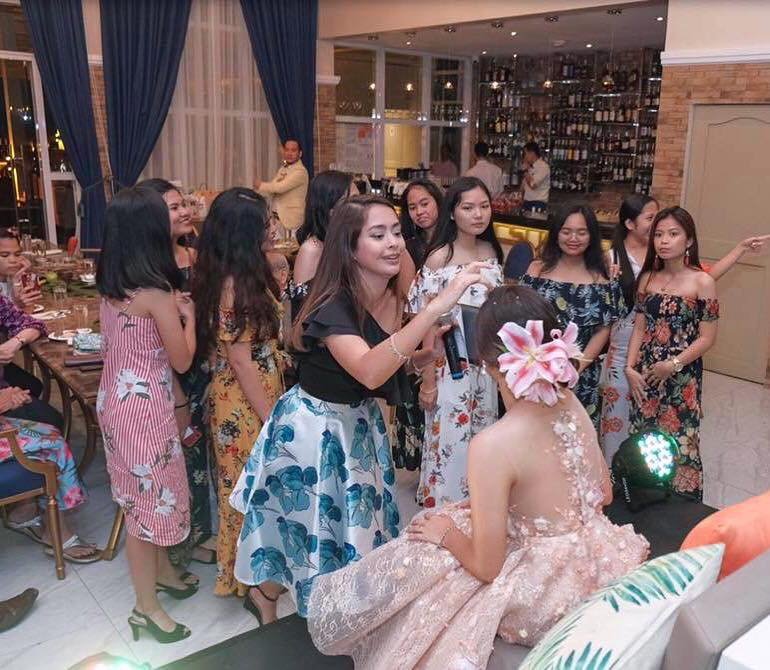 A woman in a peach dress with a flower on her head is sitting on a stage, while another woman in a black top and a blue and white skirt stands next to her, holding a microphone. Several women in floral or striped dresses are gathered around, watching and smiling at an indoor event with a bar in the background.
