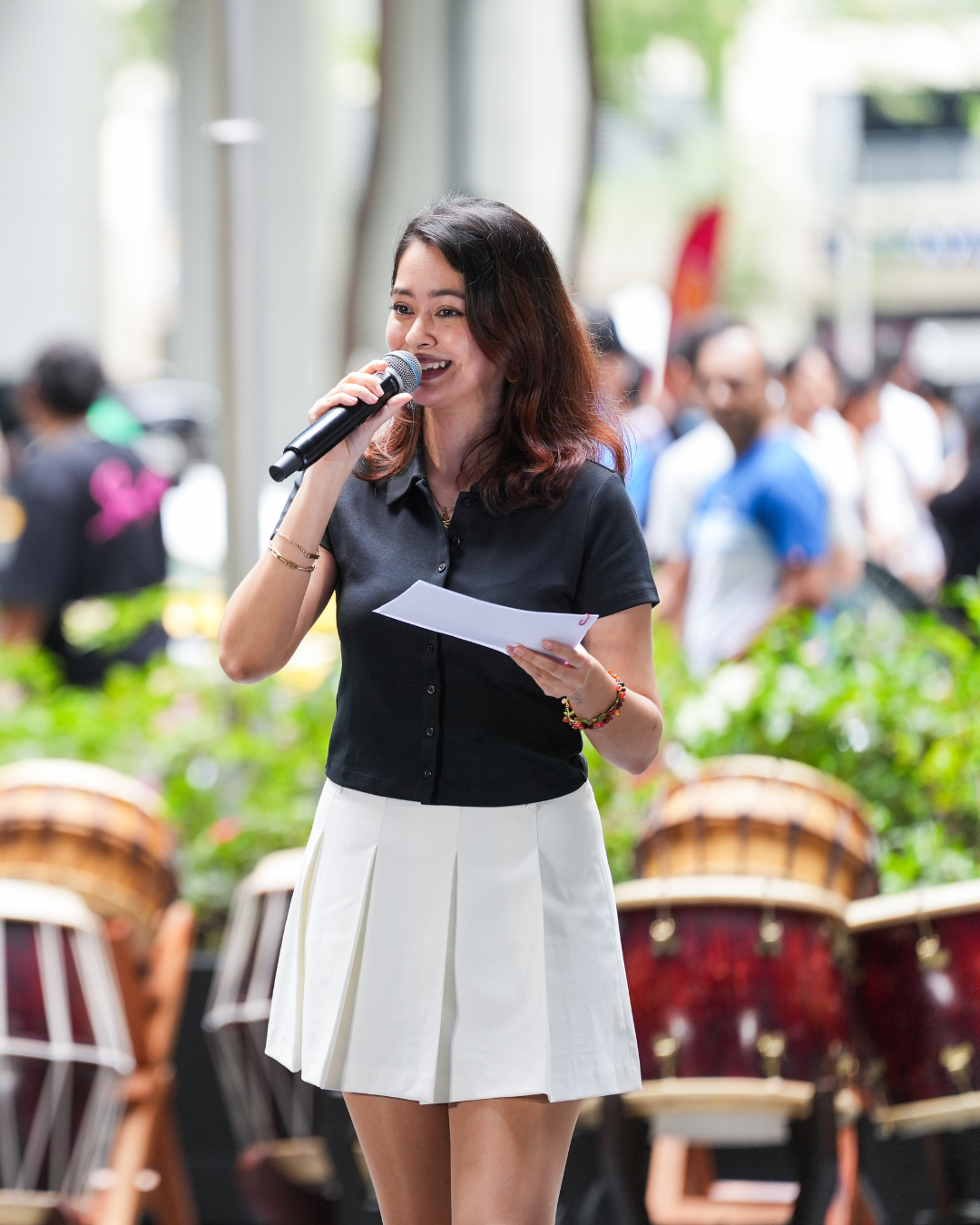 A young woman with brown hair, wearing a black short sleeved top and a white pleated skirt, speaking into a microphone at an outdoor event with a crowd and musical drums in the background.