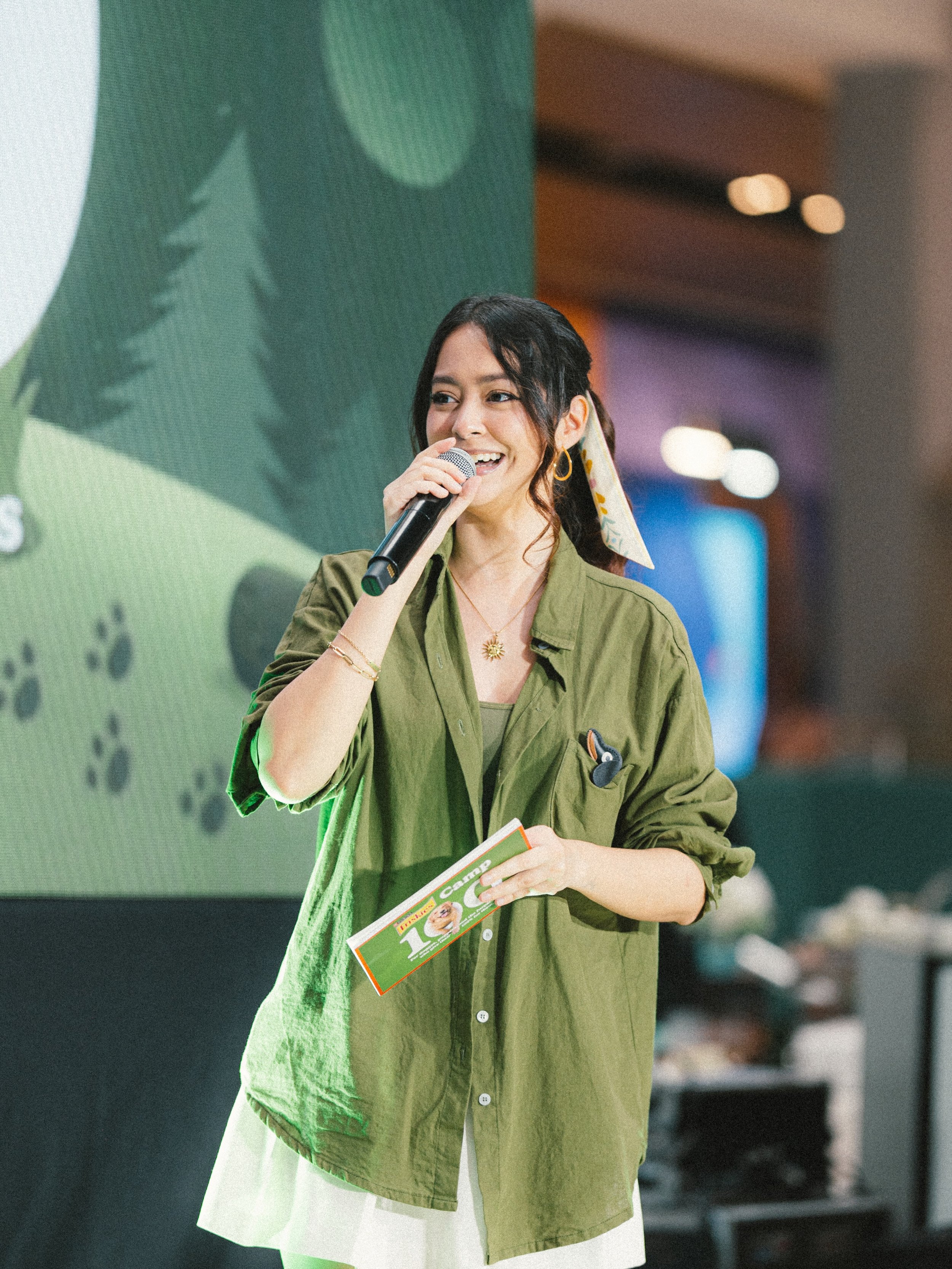 A woman smiling and speaking into a microphone, holding a pamphlet, dressed in a green shirt, standing indoors with a blurred background.
