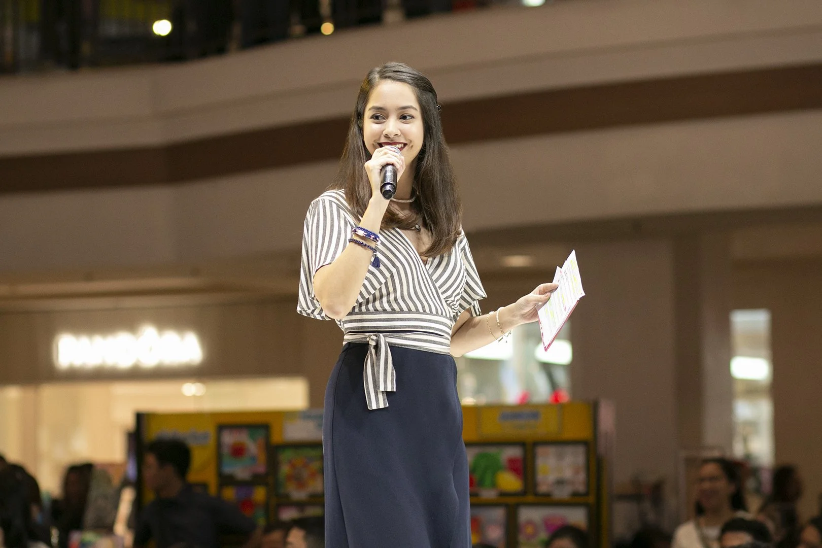 A young woman with long brown hair holding a microphone and a piece of paper, speaking in a shopping mall.