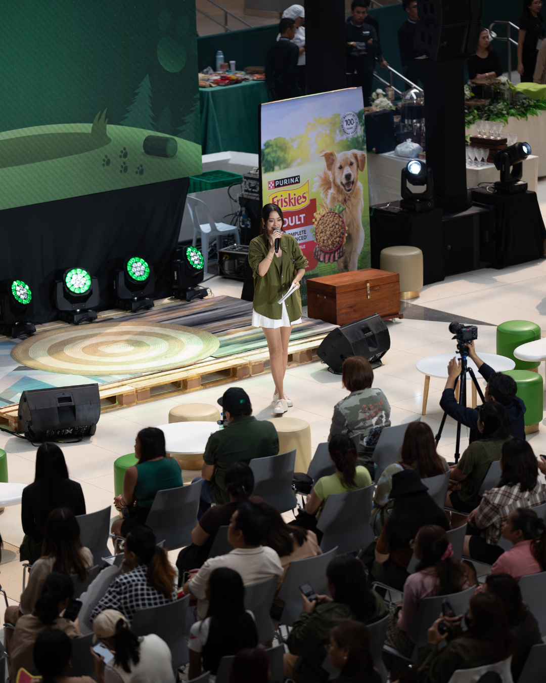 A woman on a stage speaks into a microphone at an indoor event with an audience seated in front. The event appears to be pet-related, with a large banner behind her advertising Purina Friskies dog food. The stage is decorated with a carpet, wood pallets, and lighting equipment, and a cameraman films the woman.