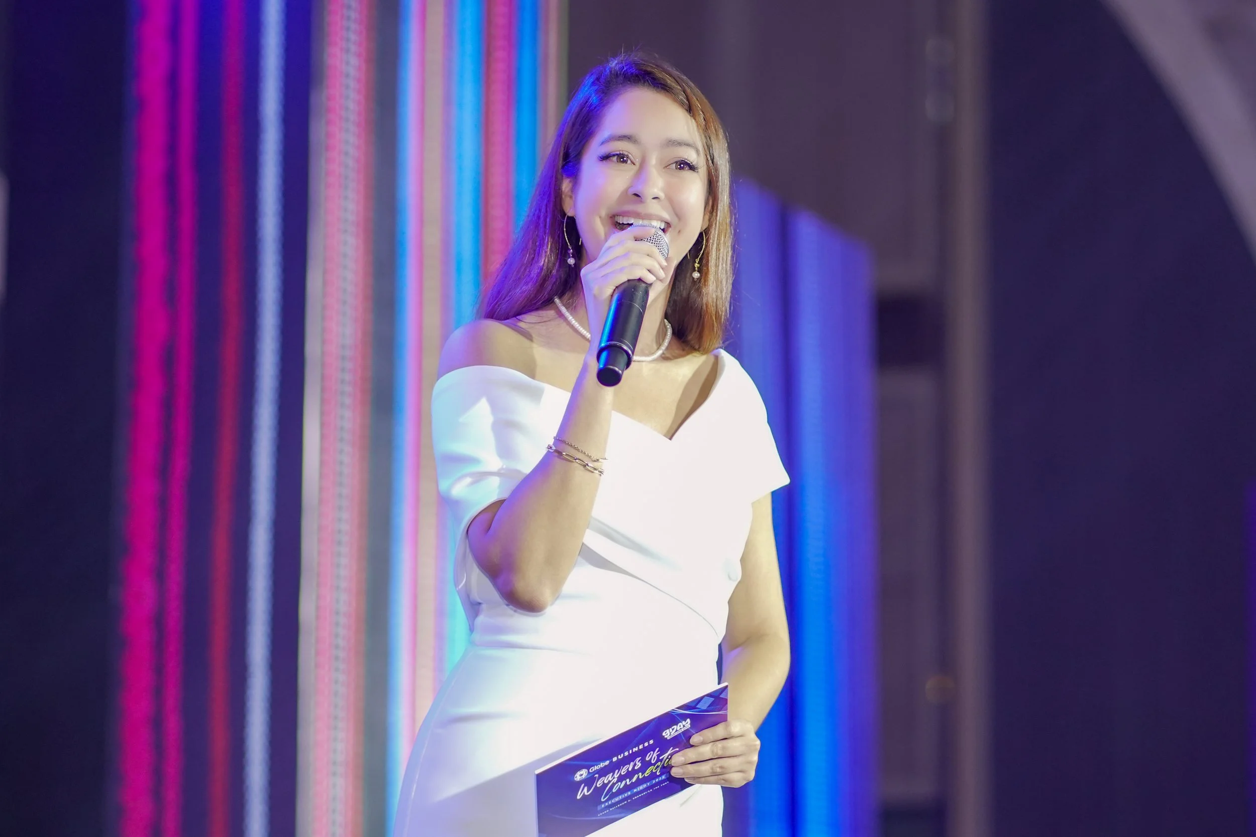 A woman with long hair and earrings speaking into a microphone at an event, wearing a white gown, with colorful vertical LED lights in the background.