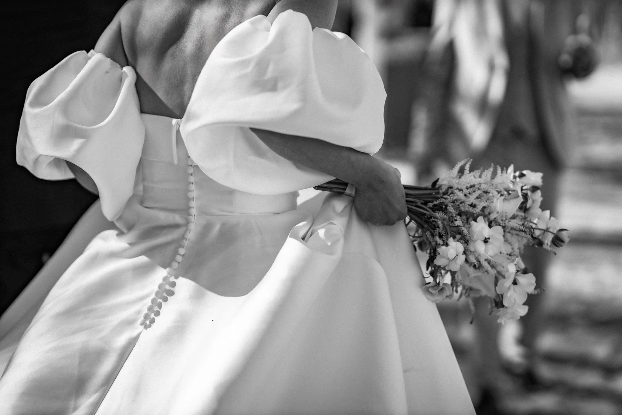 Sort-hvid foto af en person i en hvid kjole, der holder en blomsterbuket. Personen har en hånd med knyttede fingre, og der er blomsterranker i baggrunden.