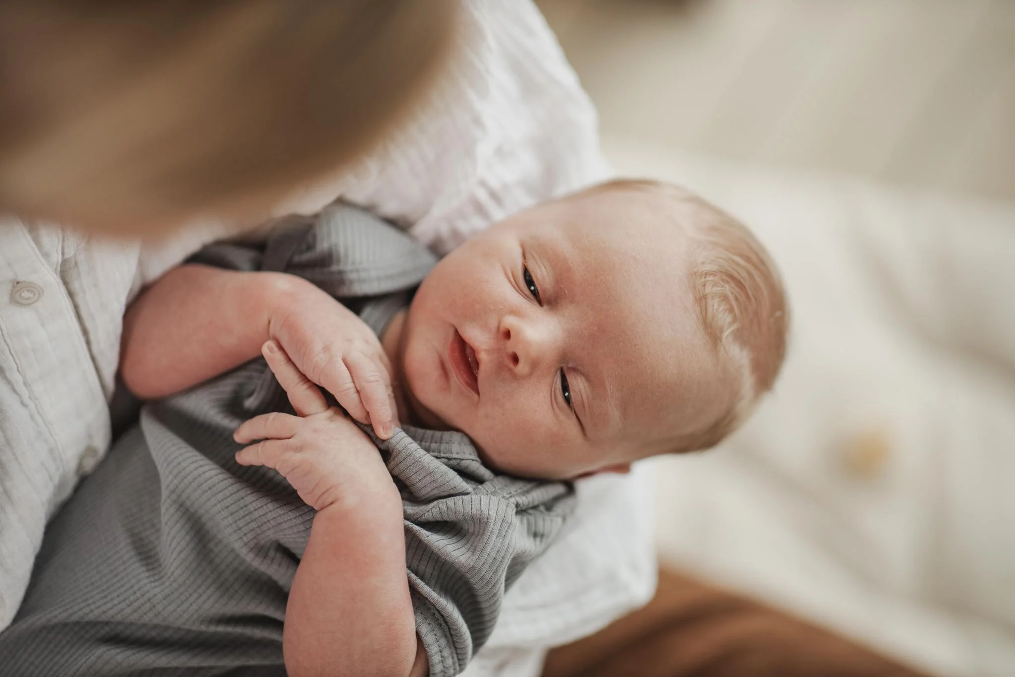 En baby bliver holdt af en voksen, smiler let og holder begge hænder op til ansigtet.