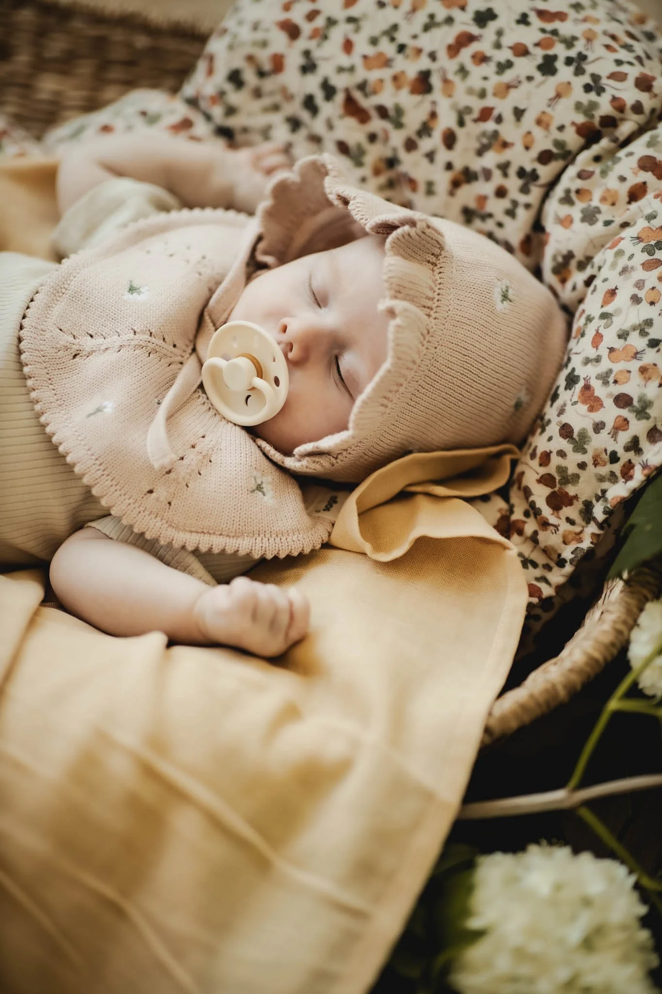 En sovende baby med pacifier, iført beige hue og sweater, ligger på en pude med blomsterprint, omgivet af blomster og grønne blade.